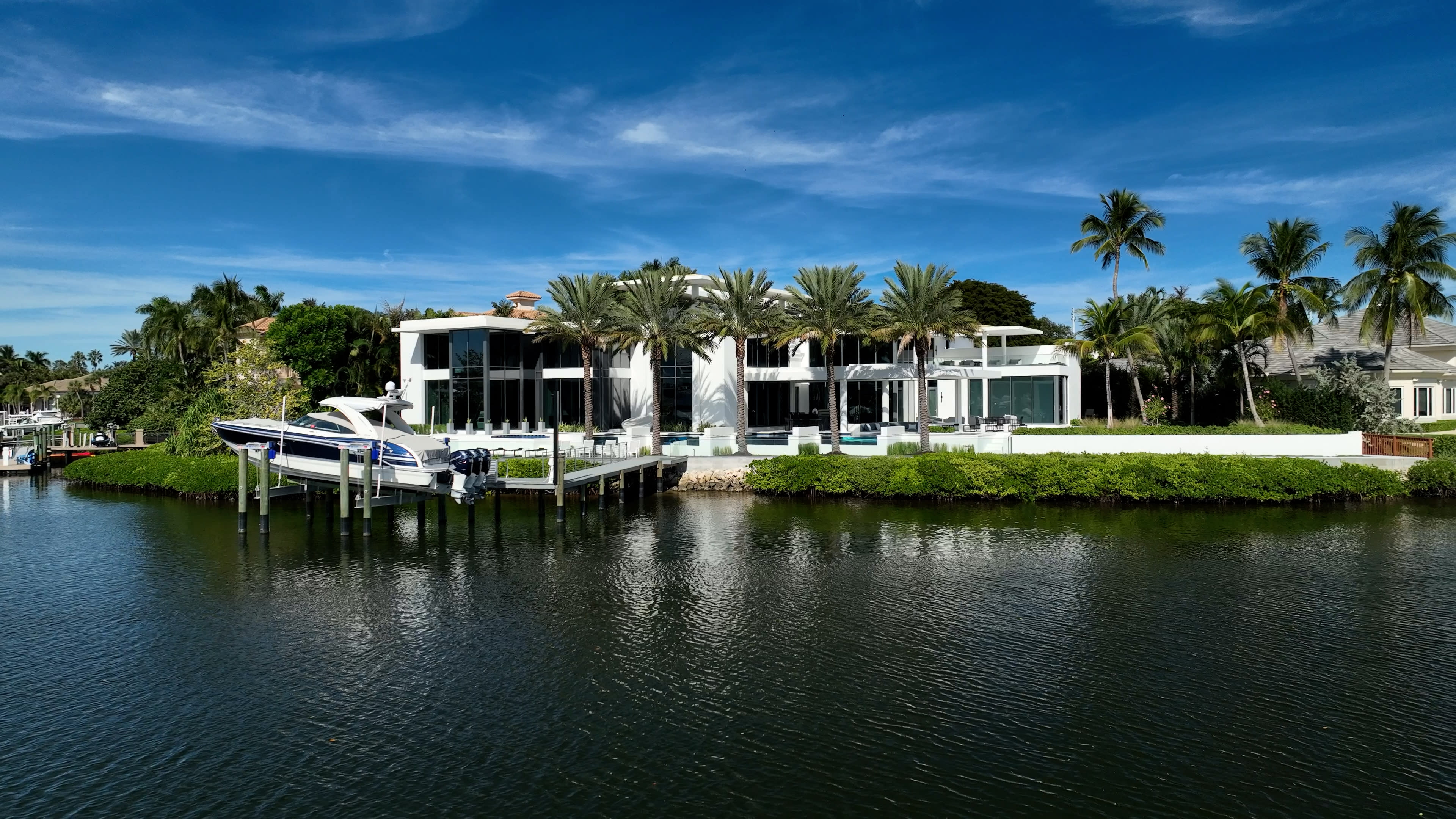 Luxurious waterfront mansion with modern architecture, surrounded by palm trees and lush greenery. A yacht is docked at a private pier, under a clear blue sky.