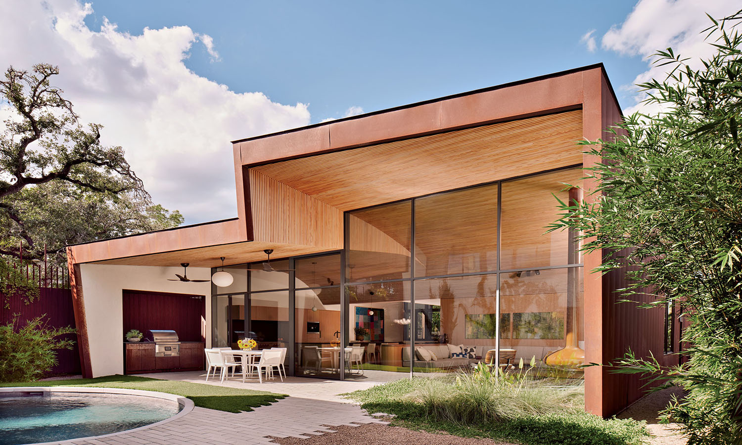 Modern house with large glass windows, wooden accents, and a covered patio with a dining set. A pool and lush green foliage surround the serene setting.