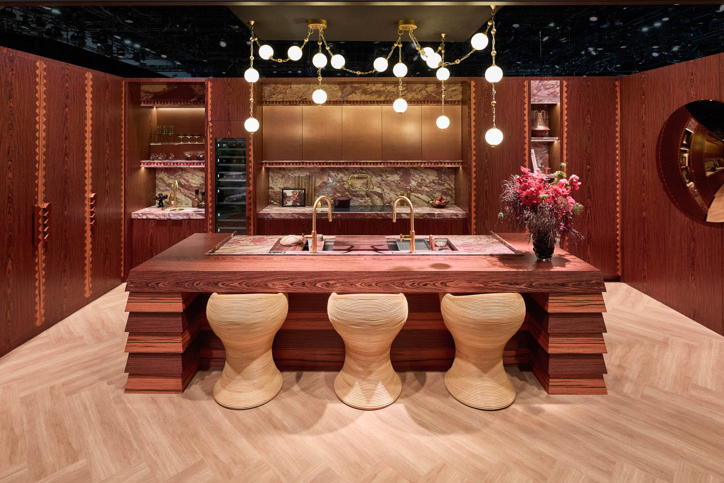 Luxurious kitchen with rich wood textures, a large island with sculptural stools, gold fixtures, globe lights, and a vibrant floral arrangement. Warm, elegant tone.