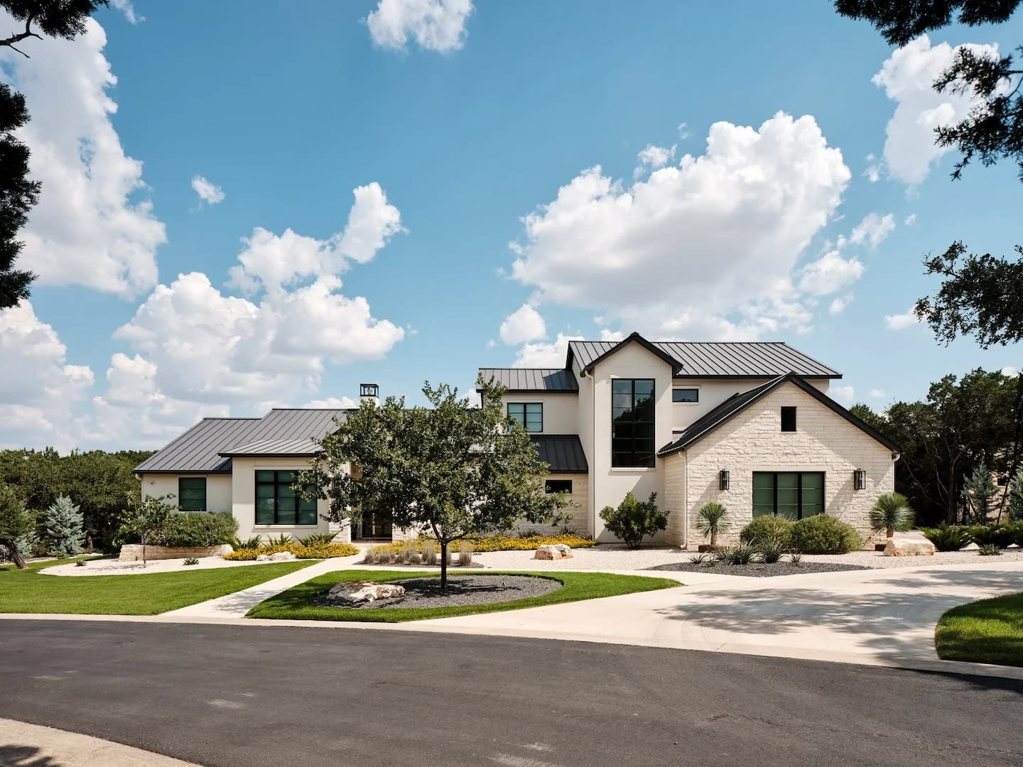 Modern suburban house with light stone exterior, dark metal roof, large windows, and landscaped yard under a bright blue sky with fluffy clouds. Calm atmosphere.