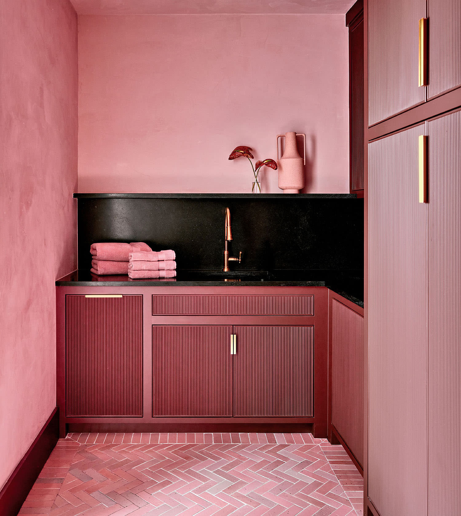 A stylish pink laundry room with textured cabinets, gold handles, and a sleek black countertop. The warm ambiance includes folded towels and decorative vases.