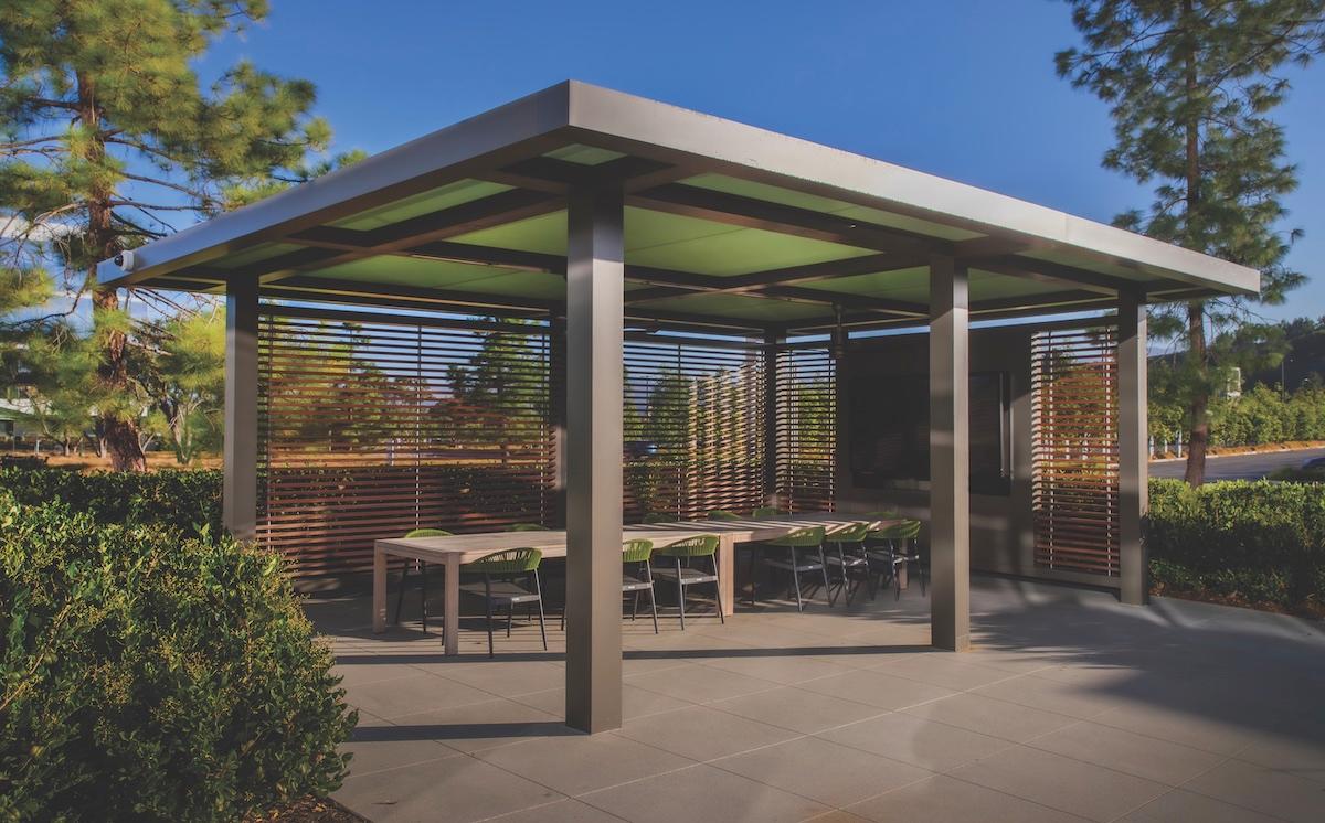 A gazebo with a table and chairs is situated in the center of a lush park, surrounded by greenery and open space.