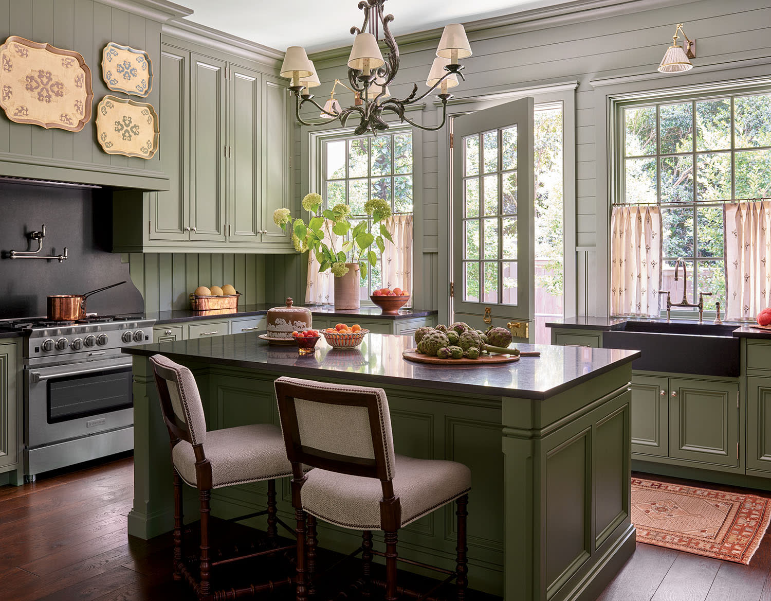 A cozy kitchen with sage green cabinets, a black countertop island, and a chandelier. Sunlight through large windows illuminates plants and fruit bowls.