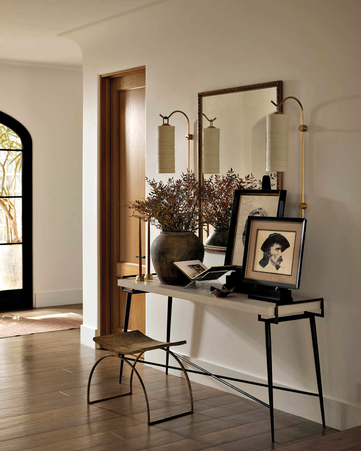 entry with a mirror, sconces, low bench and console table topped with framed art a mirror in home by Nicole Green