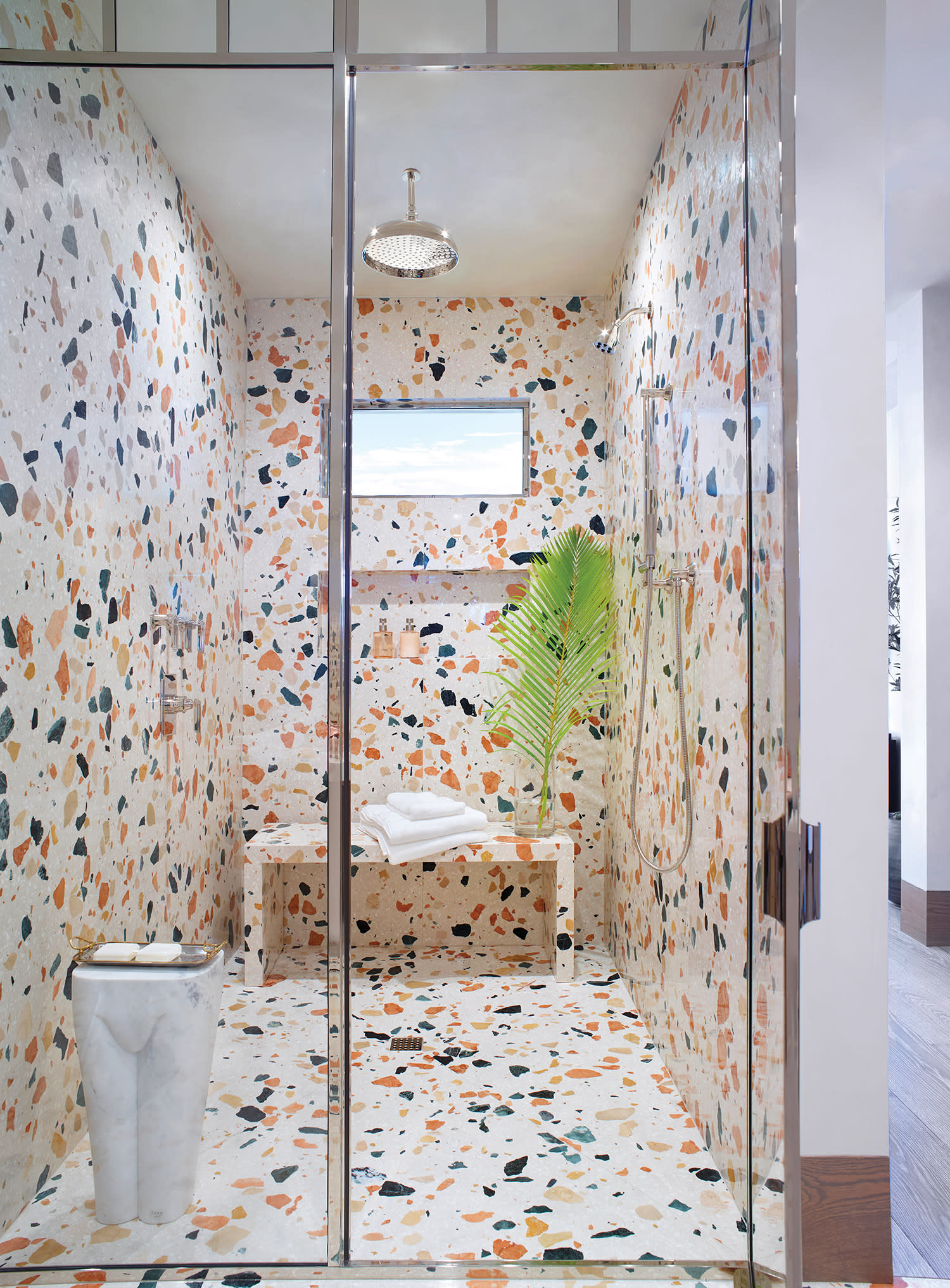 Terrazzo-tiled shower with multicolored patterns; features a chrome rain showerhead, a bench with towels, a potted palm leaf, and a small window.