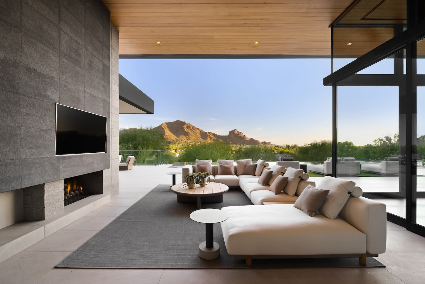 Modern outdoor living area with a large white sectional sofa, pillows, and round tables. A wall-mounted TV is above a fireplace. Mountain view at sunset.