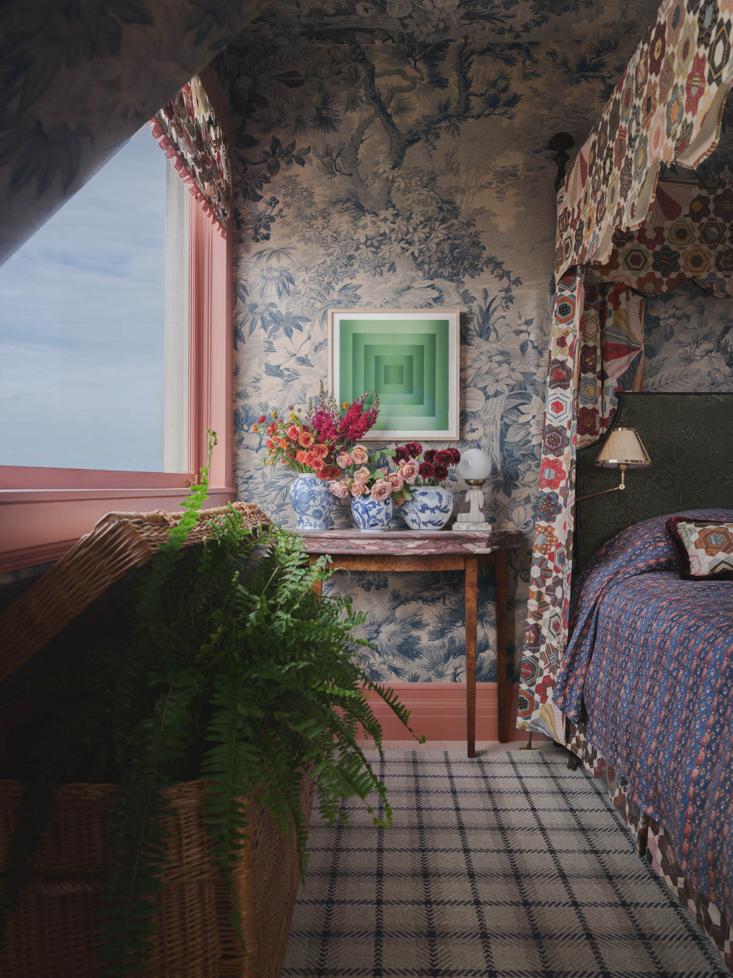 A cozy, vintage-style bedroom with floral wallpaper, plaid carpet, and a patterned bedspread. A wooden table with colorful flowers sits below abstract art.