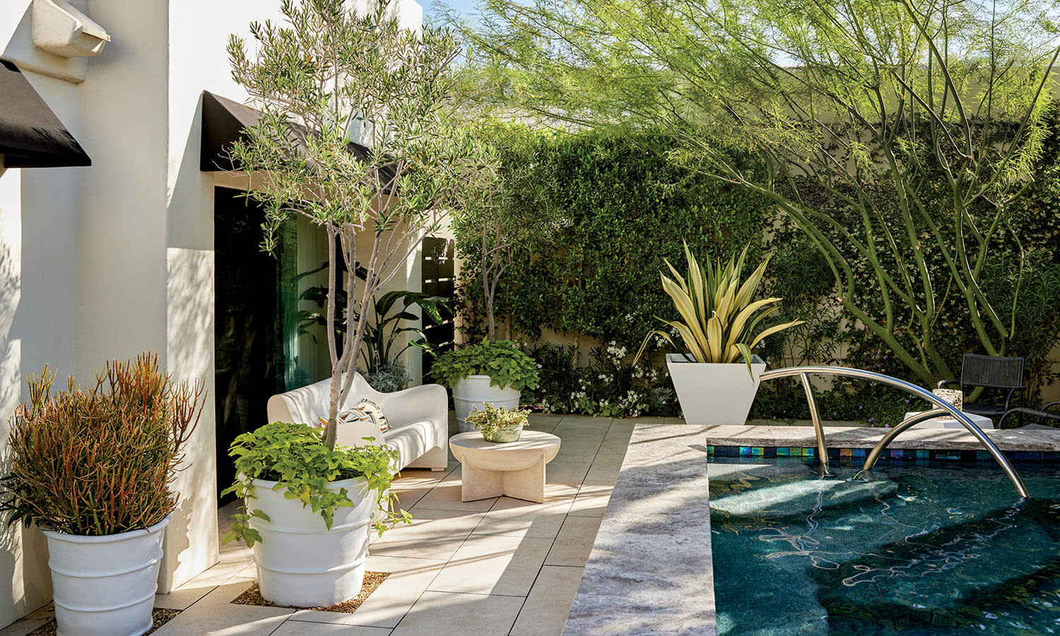 Modern patio with potted plants, white sofa, small tables, and a sparkling pool. Lush greenery surrounds, creating a serene, inviting atmosphere.