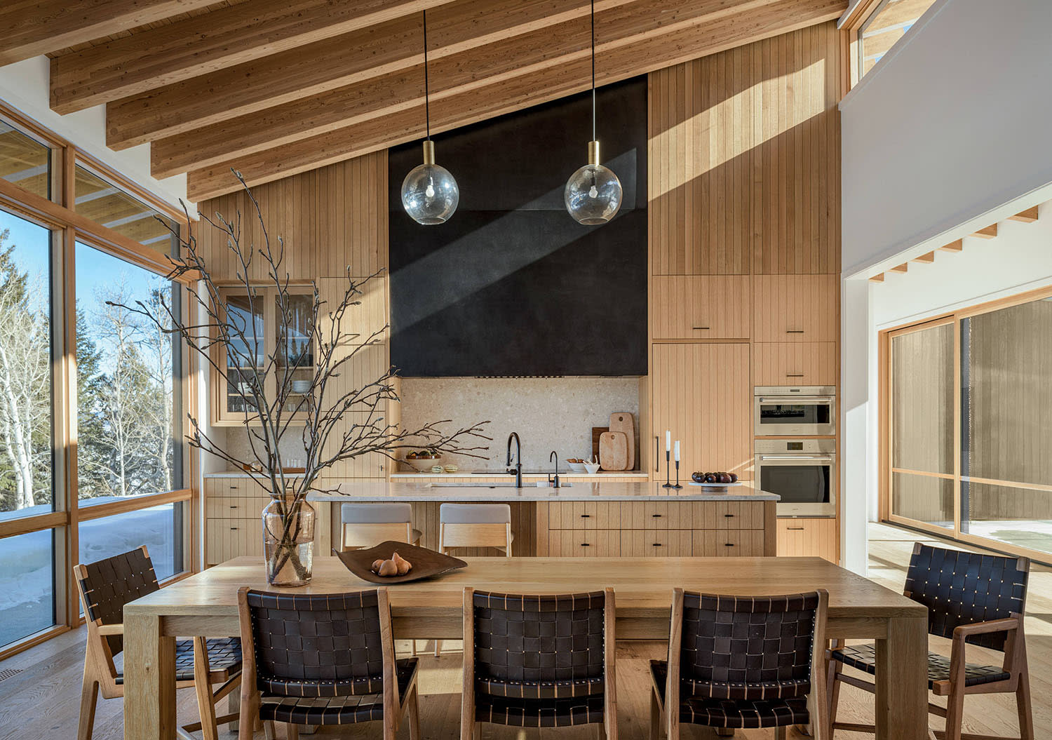 A wood dining table is surrounded by chairs in front of a kitchen area with a sloped ceiling.
