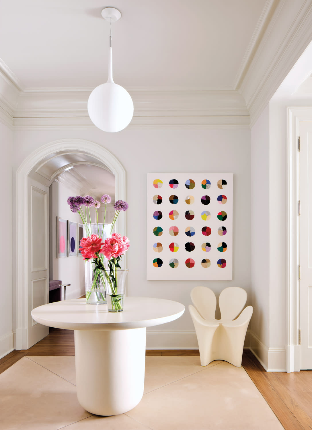 Modern white hallway with a round table holding a vase of pink and purple flowers. Colorful dot artwork on the wall and a unique cream chair.