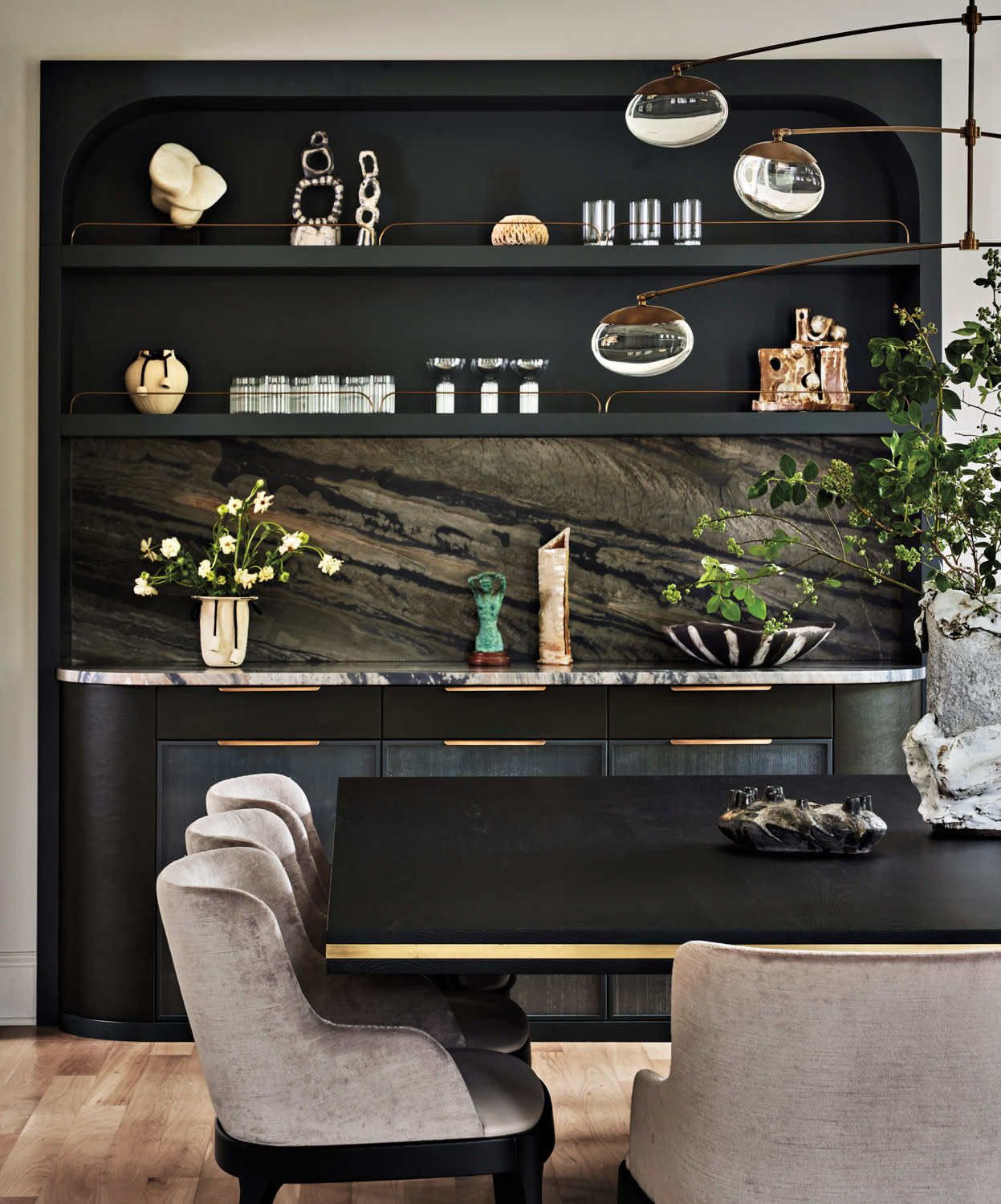 Luxurious dining room with a dark wood table, soft gray chairs, and a stylish black cabinet. Decor includes modern sculptures and vibrant greenery.