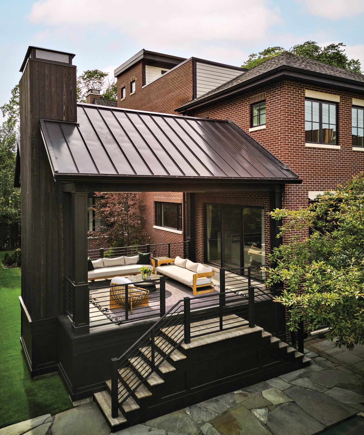 Modern brick home with a sleek black metal porch roof. Features an elevated outdoor seating area with sofas, greenery, and a serene ambiance.