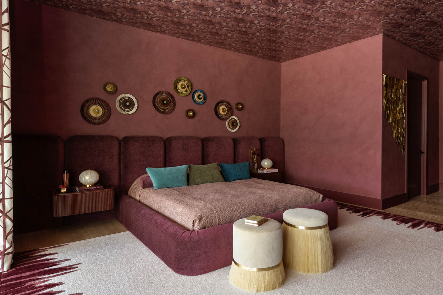 A cozy bedroom with deep burgundy walls and ceiling. A large bed with colorful cushions sits in the center, flanked by wooden side tables with lamps.
