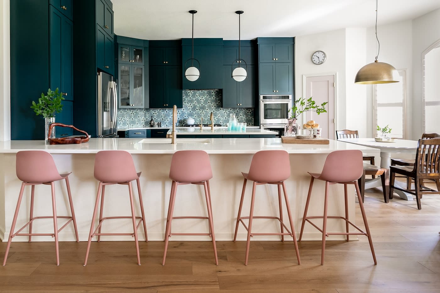 Modern kitchen with blue cabinets, patterned backsplash, and wooden floor. Five pink stools line a white island, with pendant lights above. Cozy and stylish.