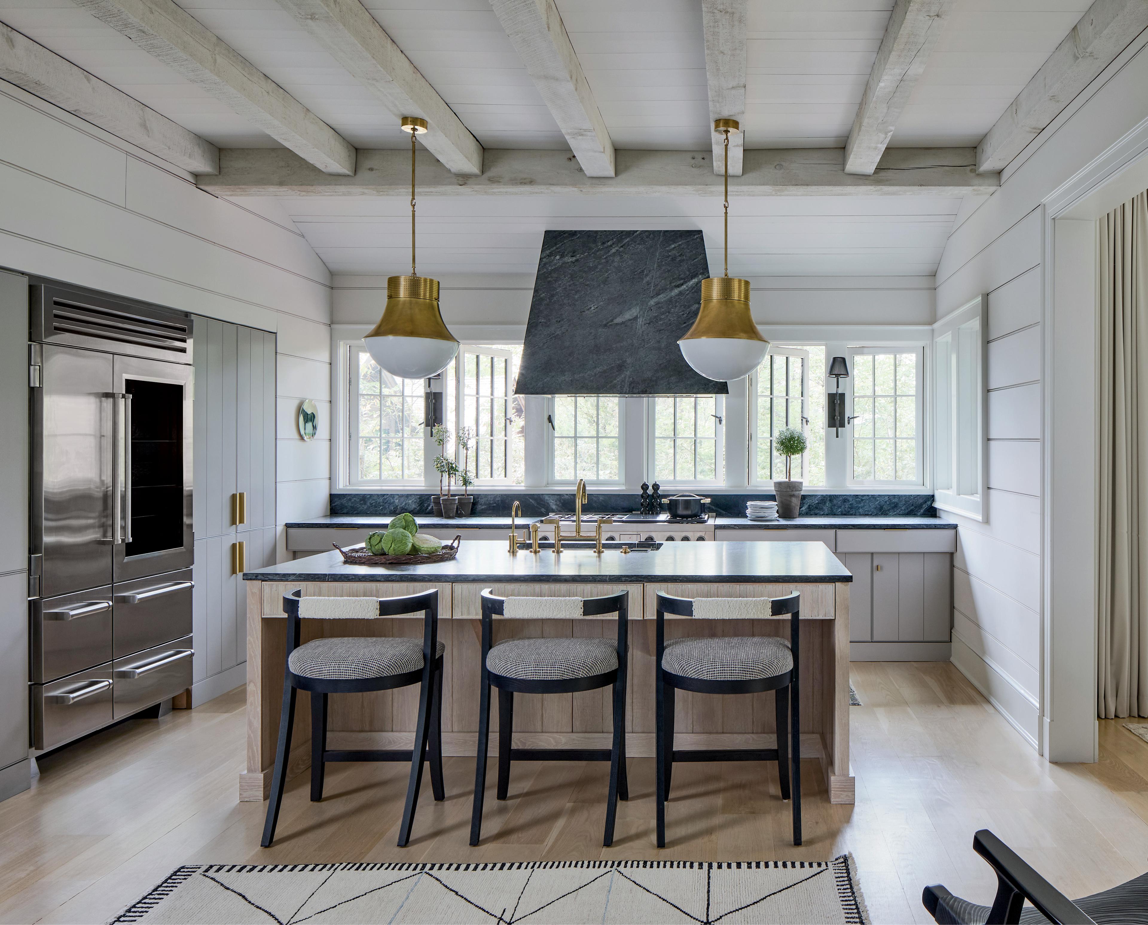 Sleek kitchen with marble island, black chairs, gold pendant lights, and stainless steel appliances. Bright, airy space with modern decor.