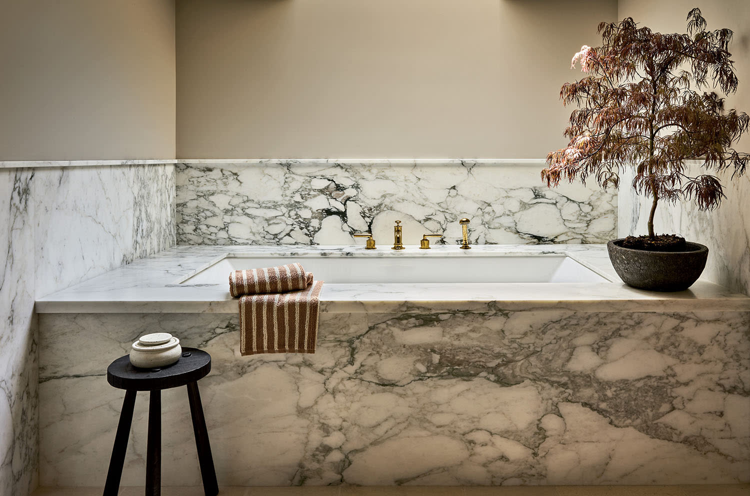 Luxurious bathroom with a marble bathtub, gold faucets, and a small tree in a pot on the edge. A striped towel and stool add warmth and elegance.