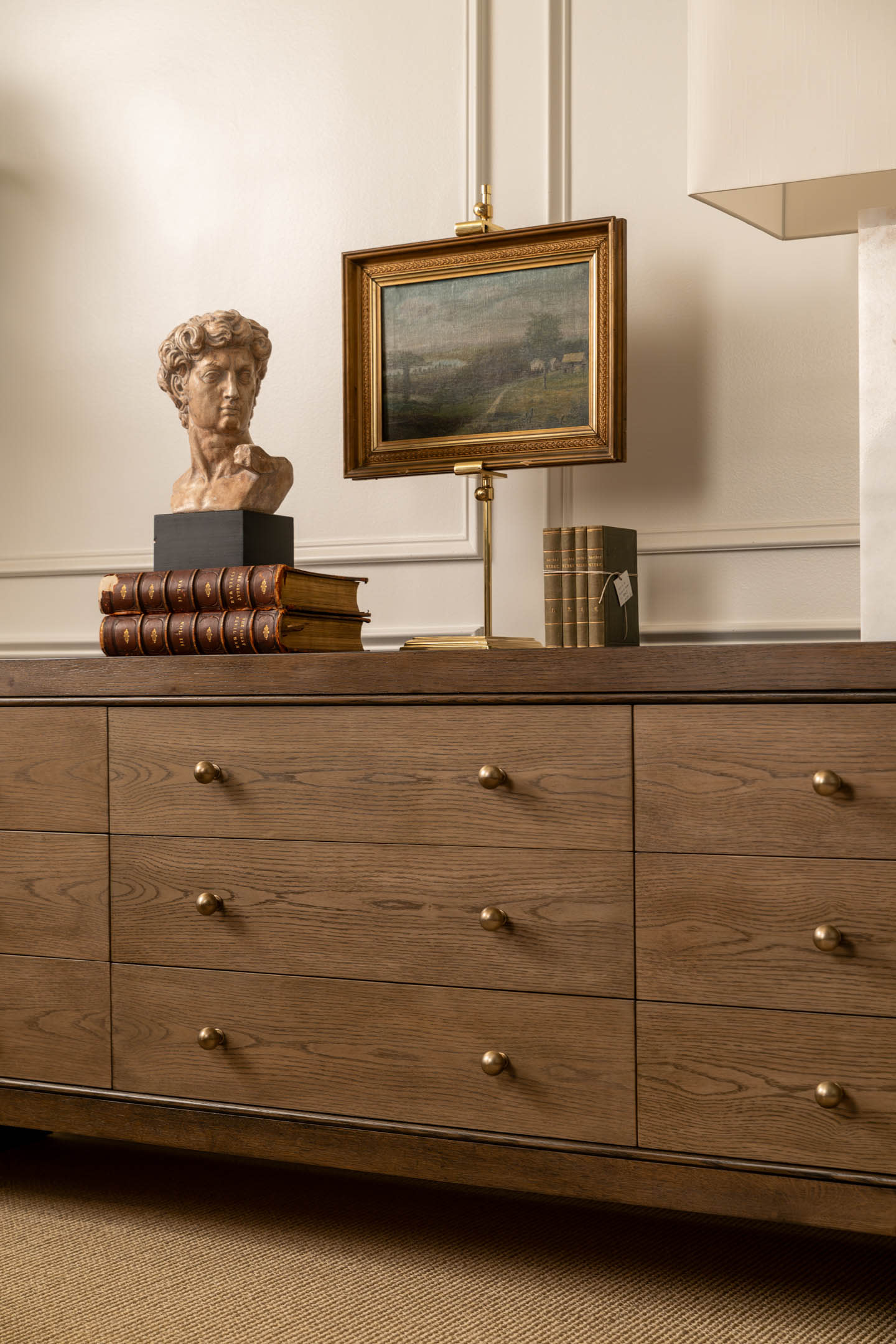 Wooden dresser with brass handles, adorned with a marble bust, stacked leather-bound books, a framed painting, and a lamp, set against a cream wall.