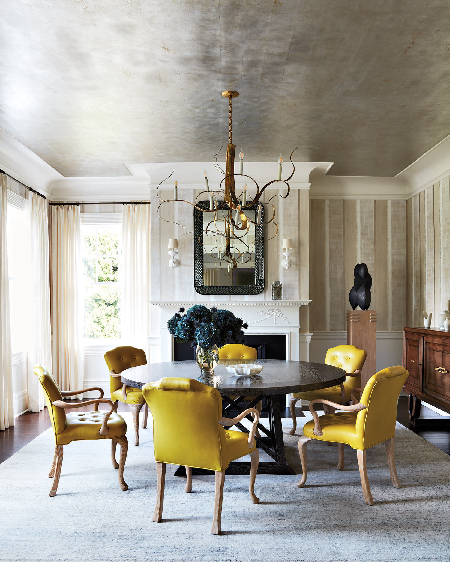 Sophisticated dining room with round table and six vibrant yellow chairs. Elegant chandelier, large mirror, and soft natural light create a refined ambiance.