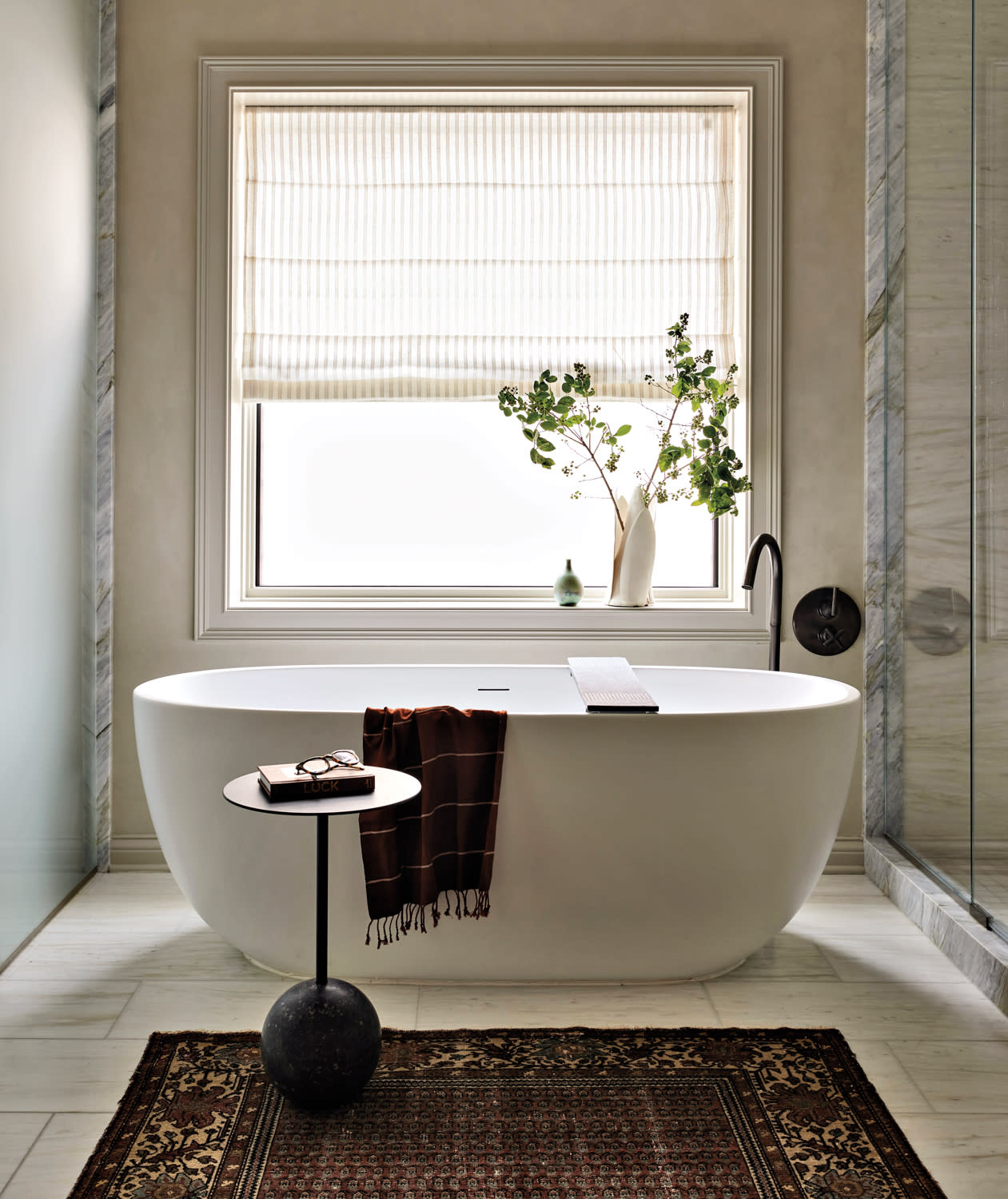 Luxurious bathroom with a white freestanding tub, vase of green branches on the windowsill, soft beige tones, cozy carpet, and a minimalist side table.