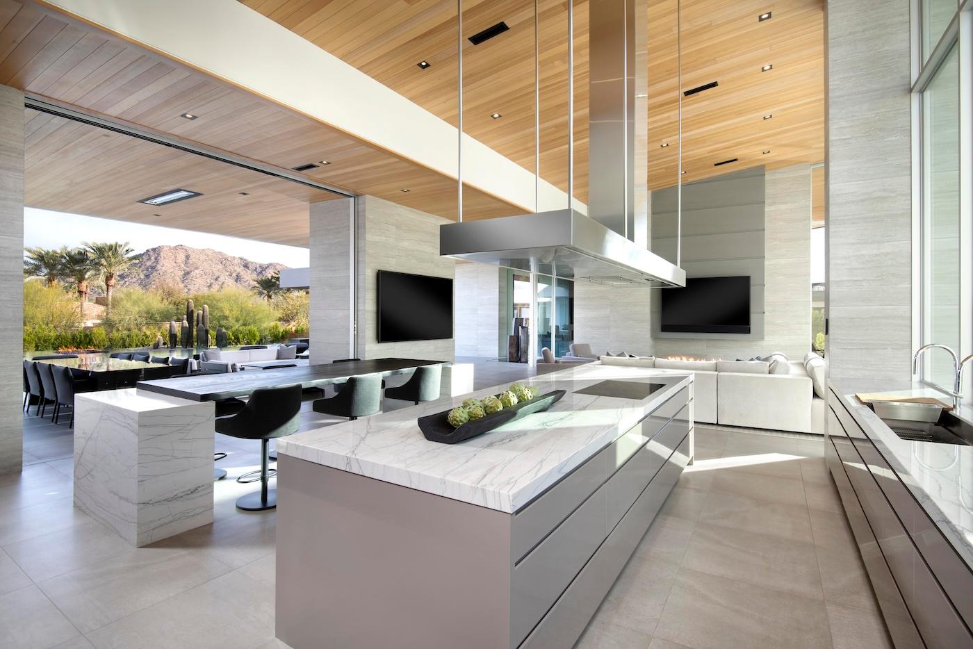 Modern kitchen and living area with sleek marble countertops, large windows, and a view of mountains. Open, airy space with a contemporary vibe.