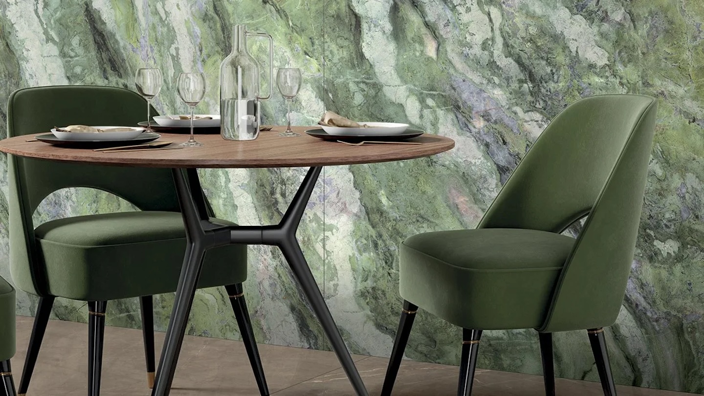 A round wooden table with a clear bottle and four wine glasses sits between two green upholstered chairs against a marble-patterned green wall.