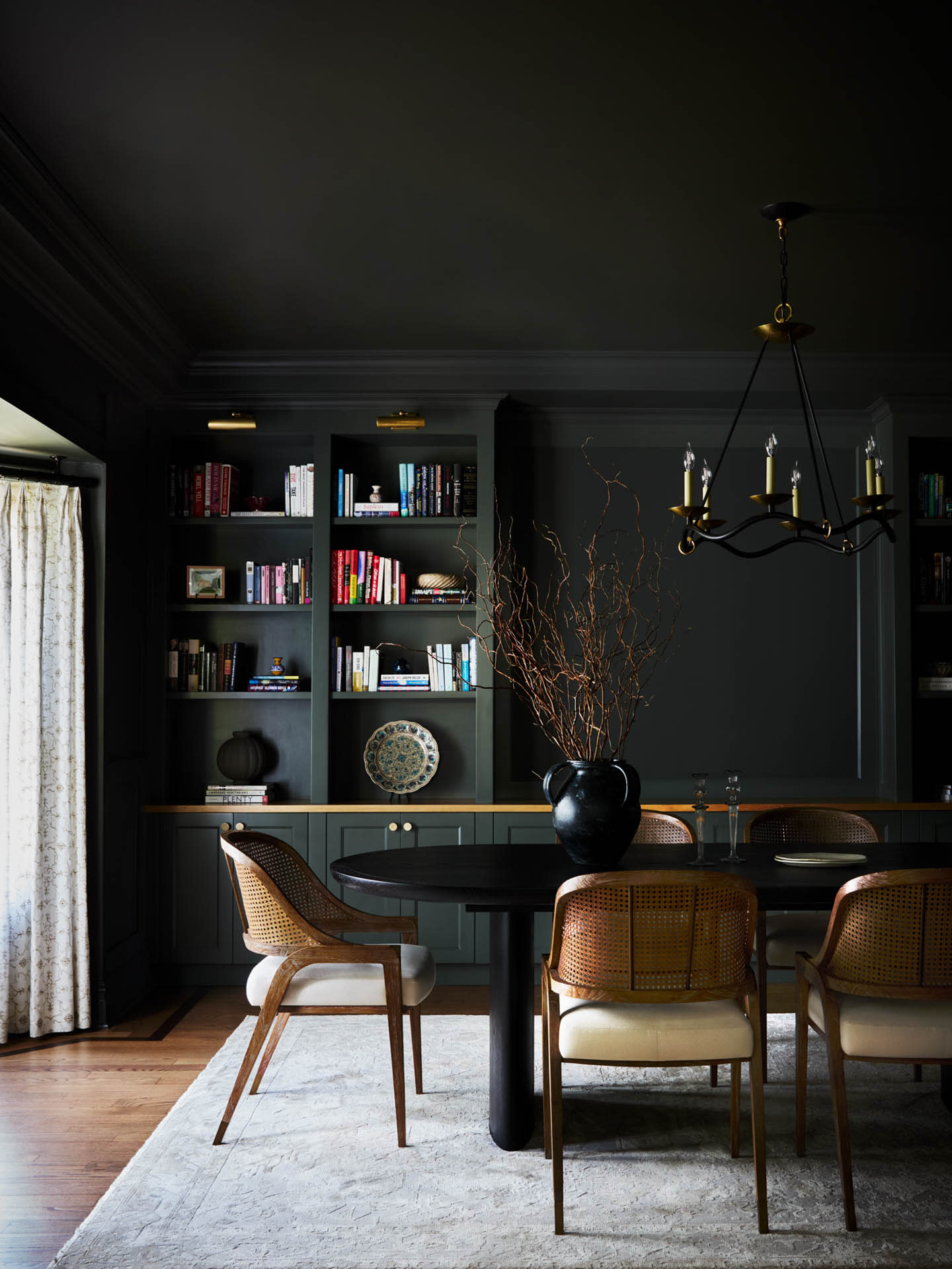 Sophisticated dining room with dark walls, built-in bookshelves filled with colorful books, a round black table, wicker chairs, and a hanging gold chandelier.