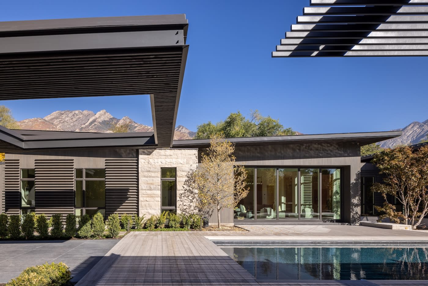 Modern house with minimalist architecture, large glass windows, surrounded by trees. A pool reflects the clear blue sky and distant mountains. Serene vibe.