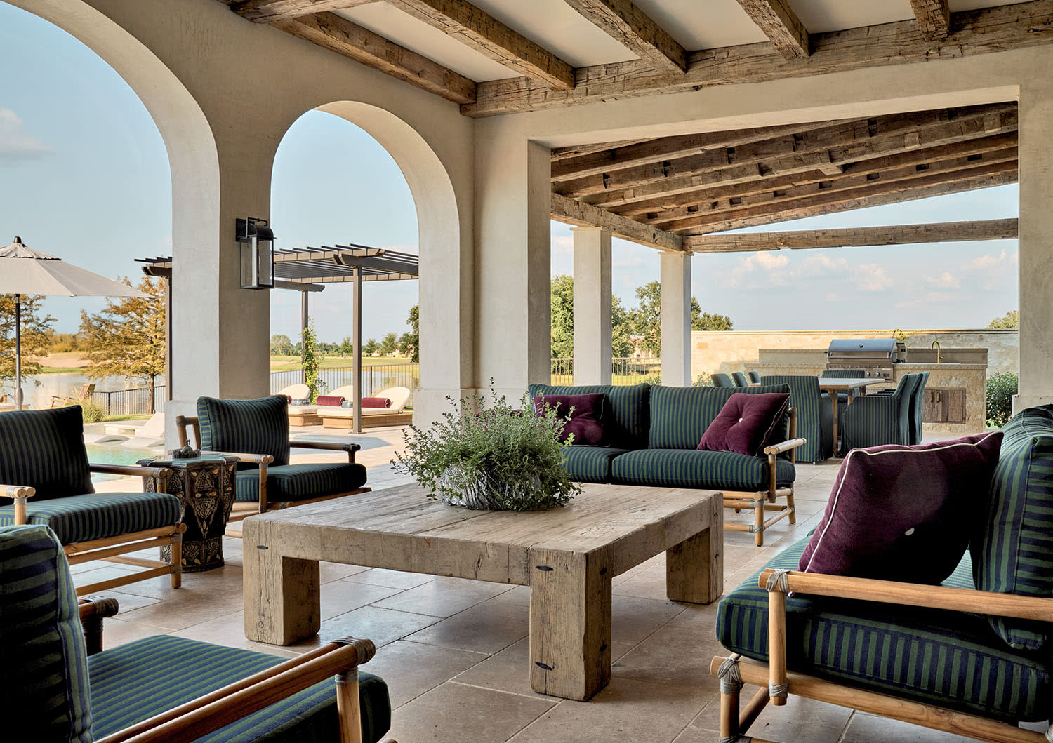 Spacious outdoor patio with rustic wooden beams, cushioned chairs, and a central table. Arched openings lead to a sunny pool area, creating a relaxing vibe.
