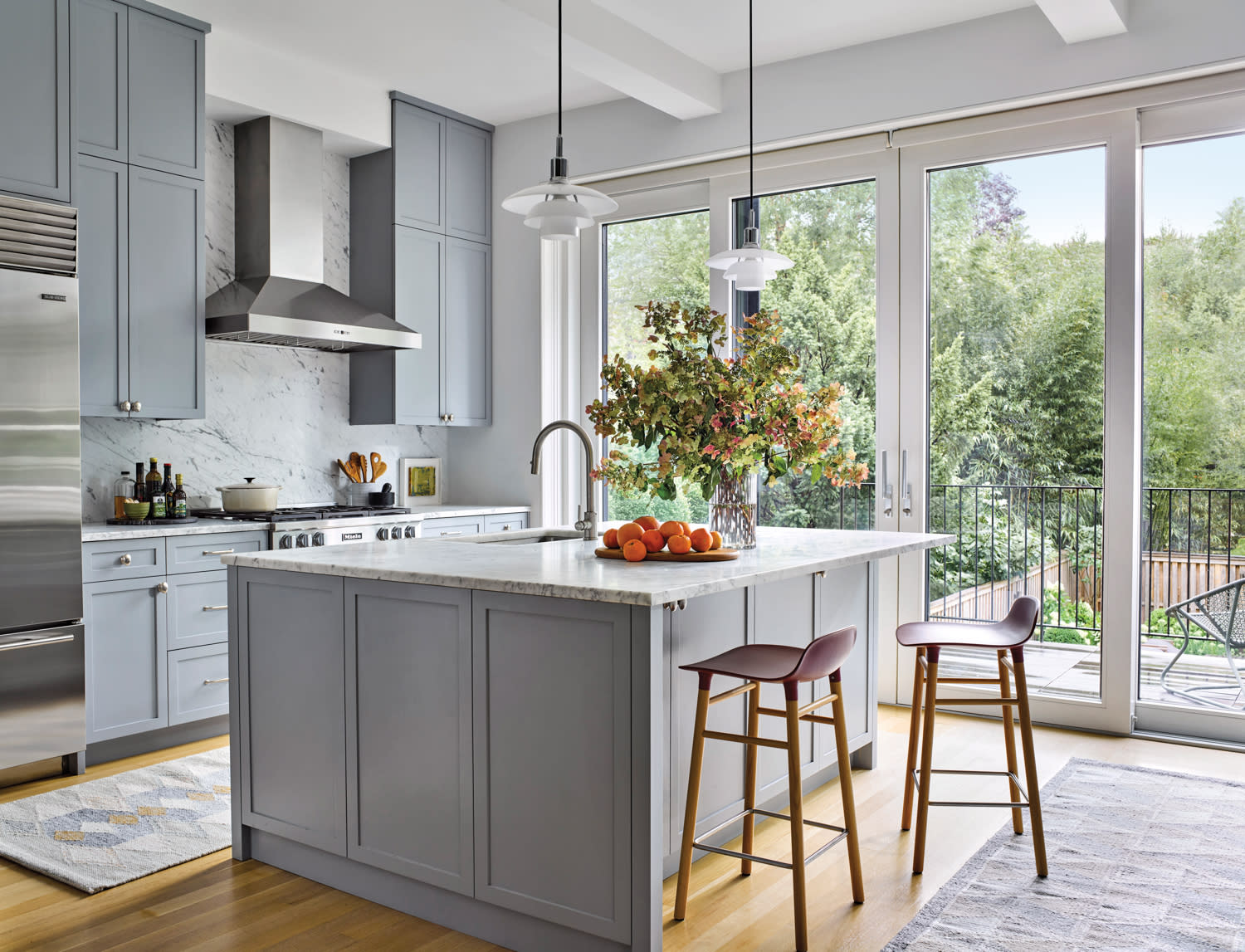 Modern kitchen with gray cabinets, marble backsplash, and island. Sunlit space includes plants, fruits on the counter, and large glass doors to a garden.