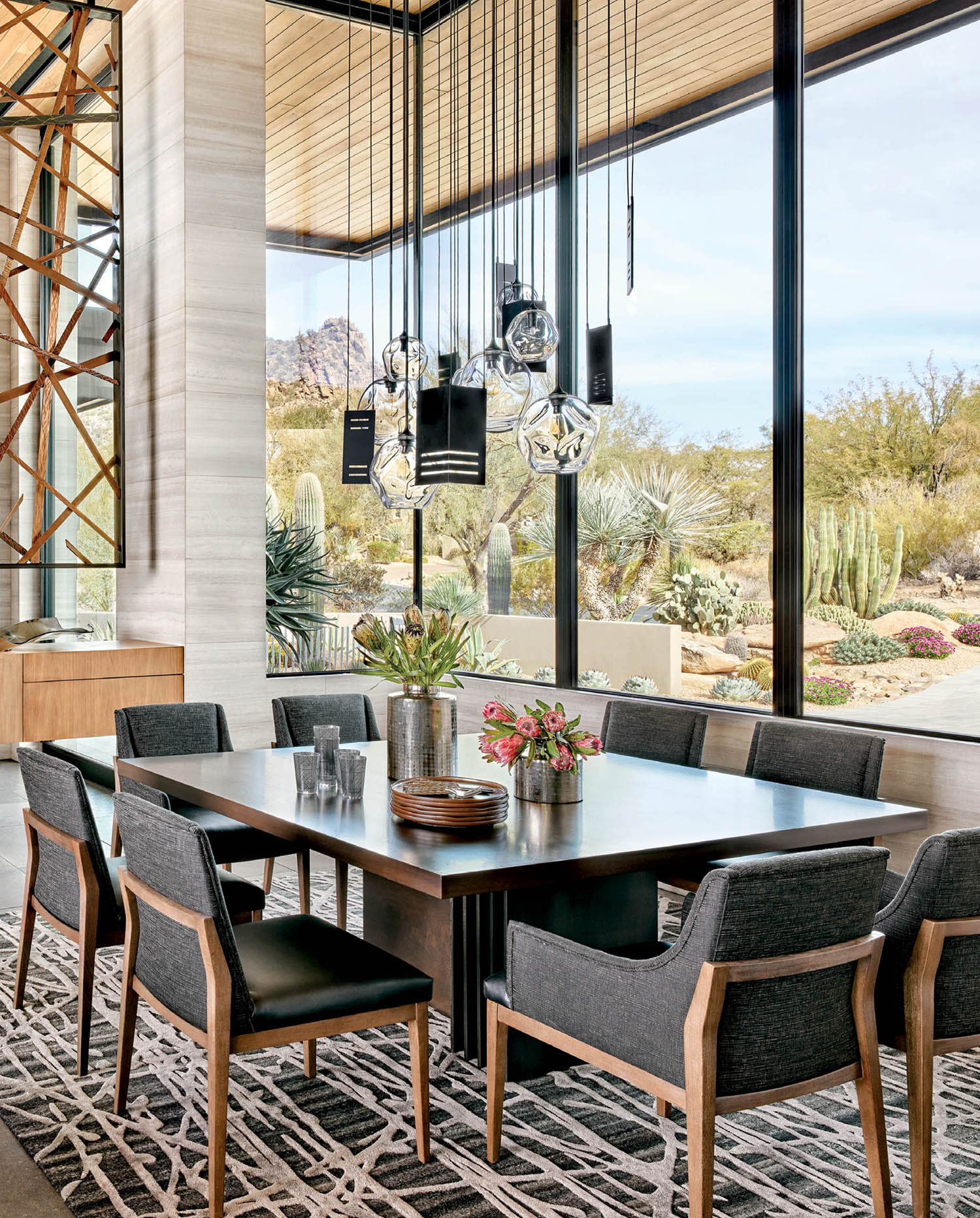 A dining room table set with chairs sits on a geometric-printed rug in front of large windows.