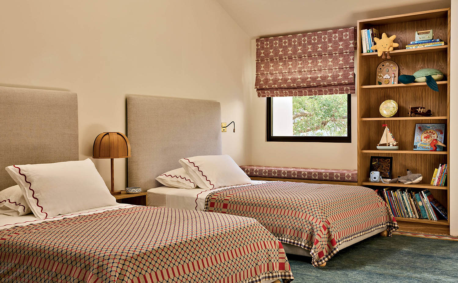 Two beds with fabric headboards and patterned quilts sit near a bookshelf holding books and toys.