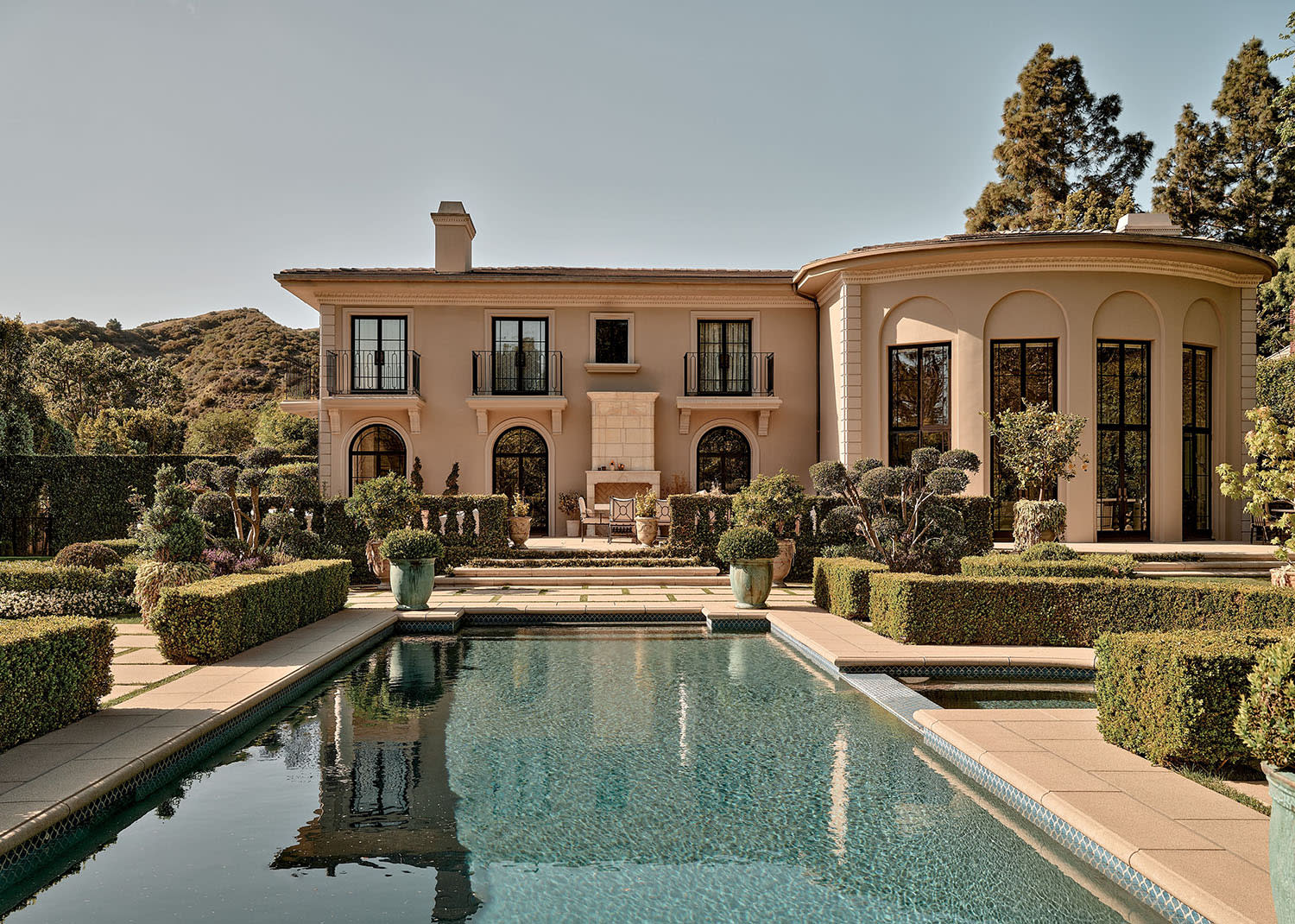Elegant beige mansion with large windows, surrounded by manicured hedges and tall trees. A serene swimming pool reflects the clear blue sky.