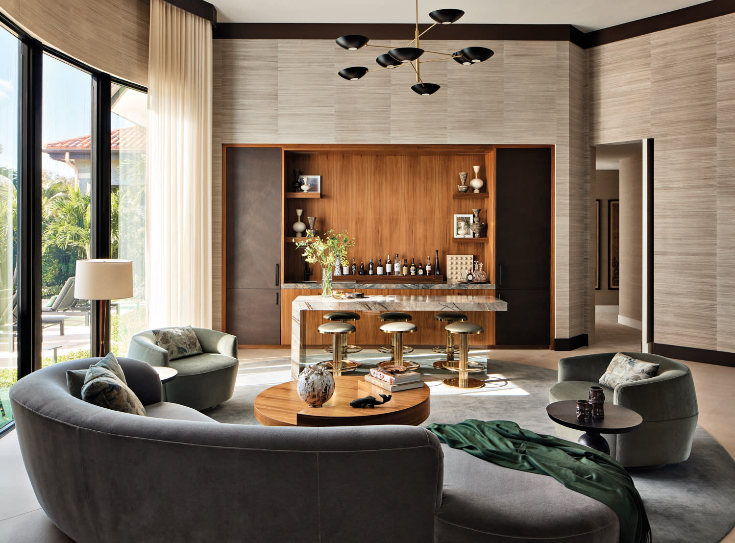 Luxurious living room with modern decor. Features a curved gray sofa, wooden coffee table, bar with stools, and large windows overlooking greenery. Elegant ambiance.