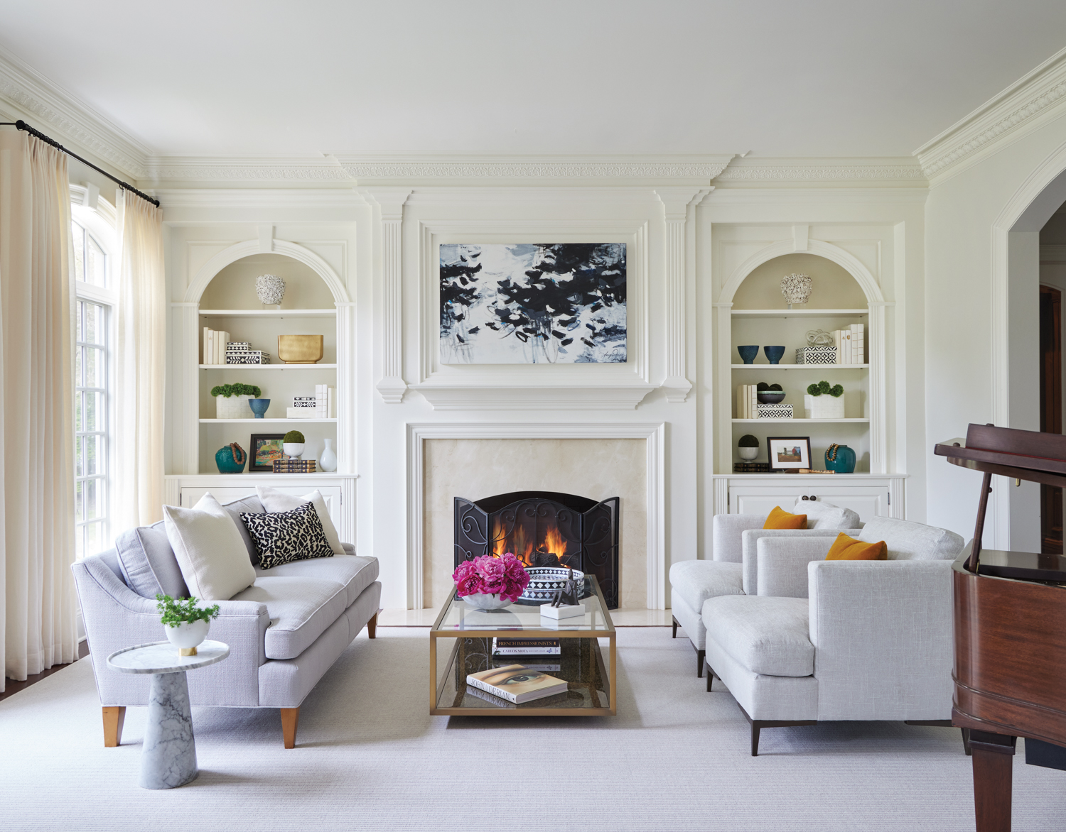 A living room with cream walls, built-in shelves, a gray sofa and a a pair of gray armchairs. Above the fireplace hangs a black-and-white abstract painting.