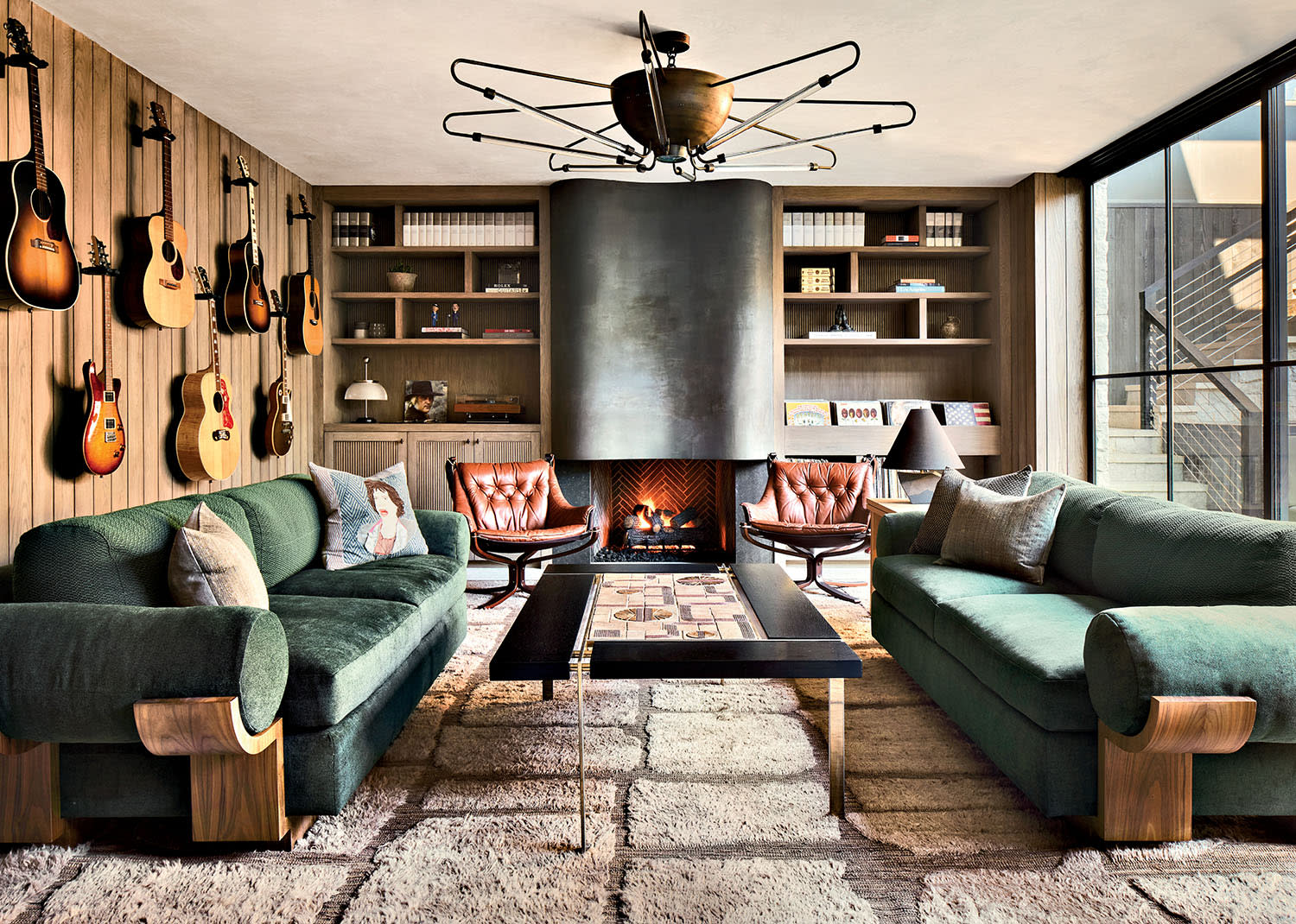 Stylish living room with green sofas, a central fireplace, and a wall adorned with guitars. Large windows and shelves create a cozy, artistic ambiance.