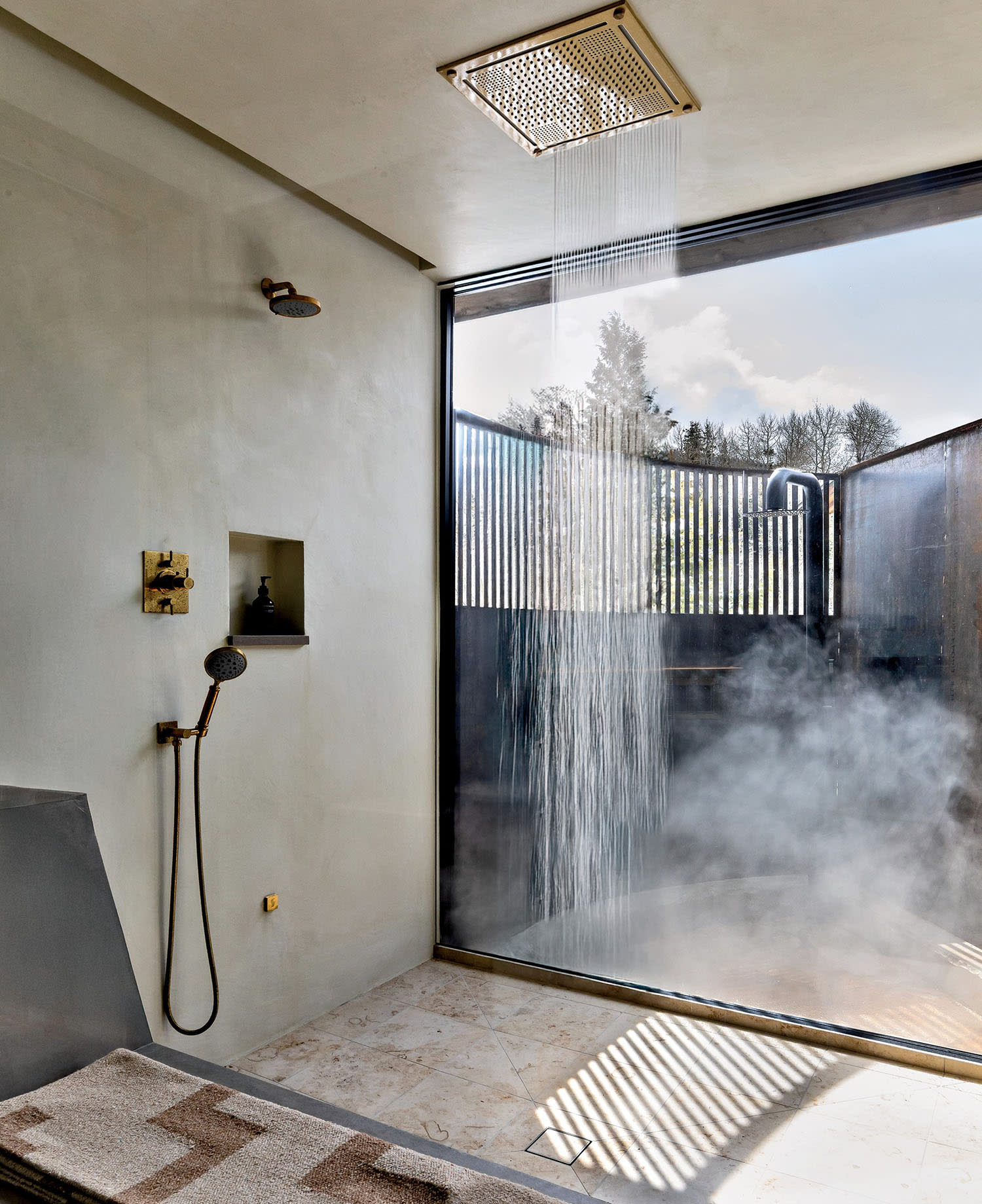 Modern shower with a waterfall showerhead streams water onto a stone floor. Sunlight and steam create a serene, spa-like atmosphere.