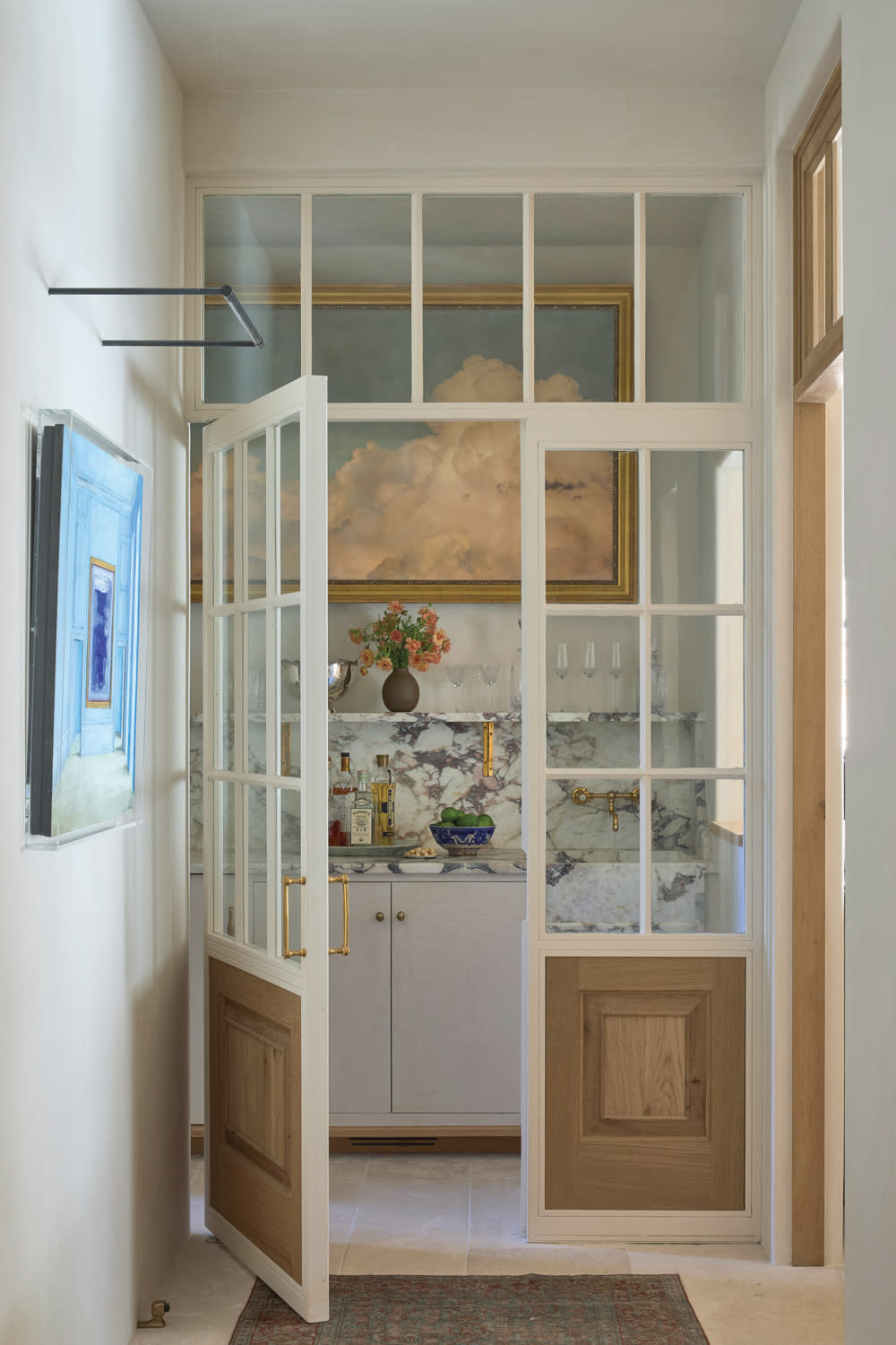 A narrow room with glass doors reveals a marble countertop, vintage-themed decor, and a large cloud painting. Soft lighting creates a cozy atmosphere.