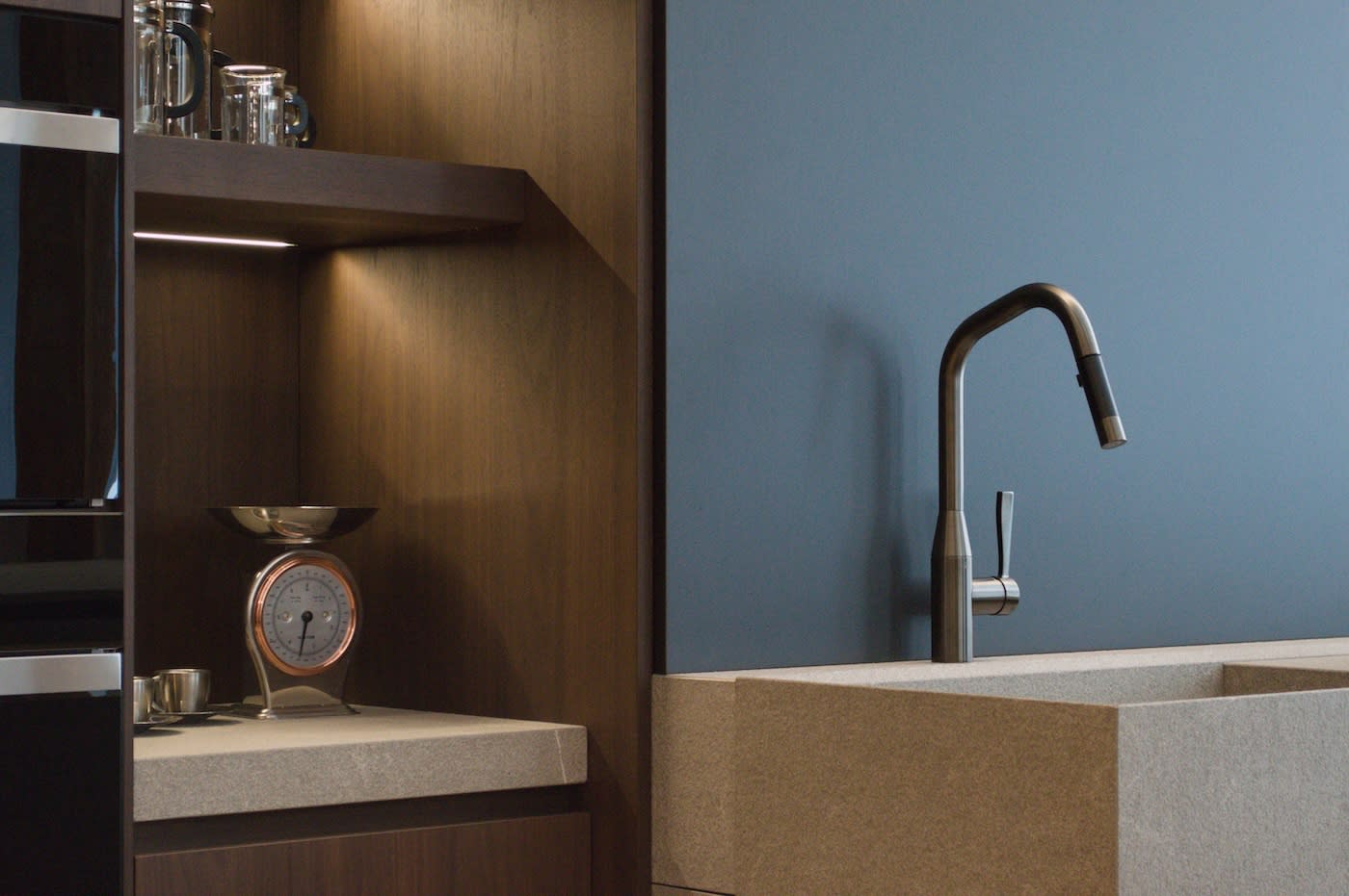 Modern kitchen with a sleek silver faucet and stone sink against a gray wall. Wooden shelves host a retro scale and glass containers, exuding a minimalist vibe.