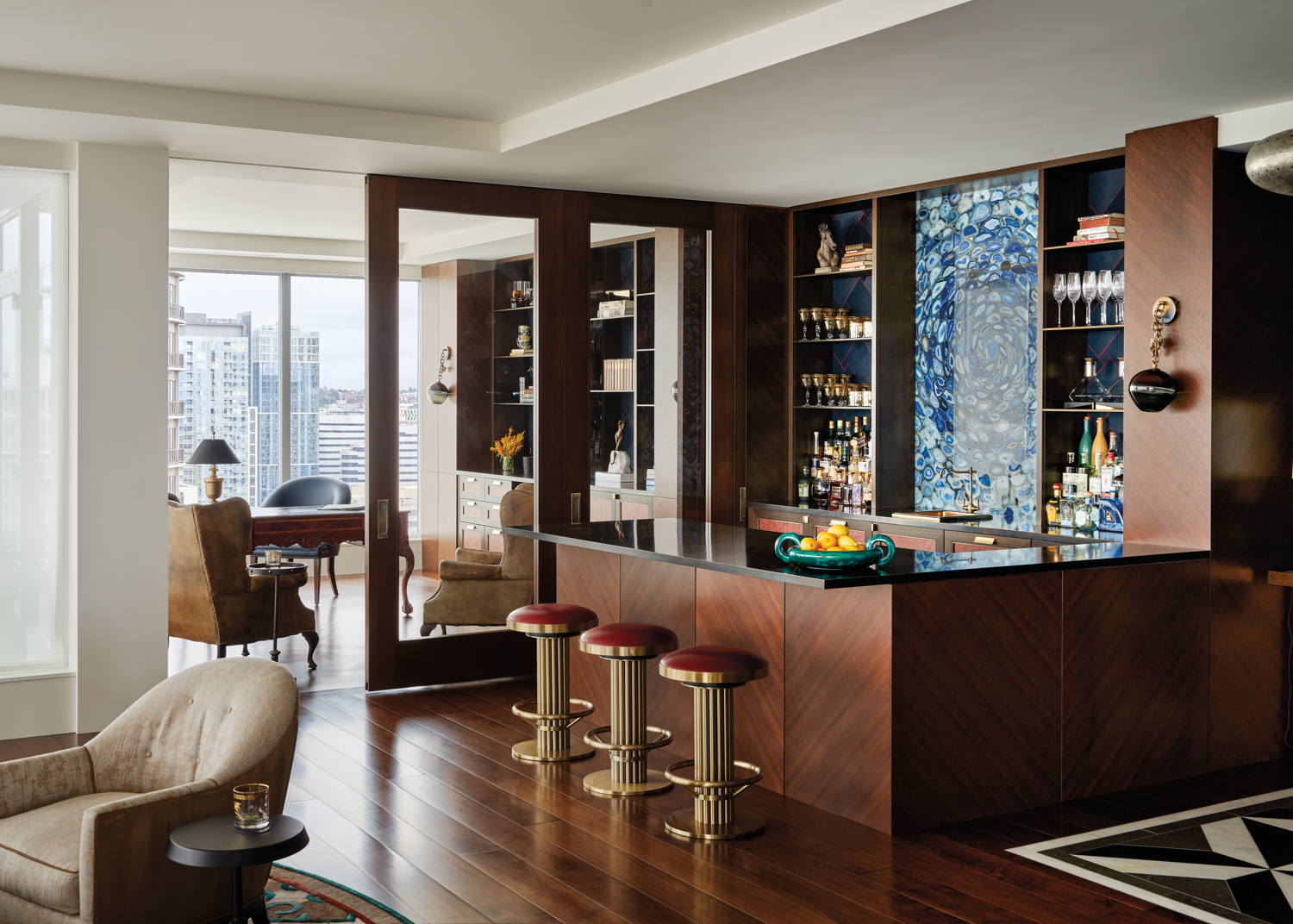 The bar area has a blue agate backsplash, upholstered stools and brass countertop in home by James Fung