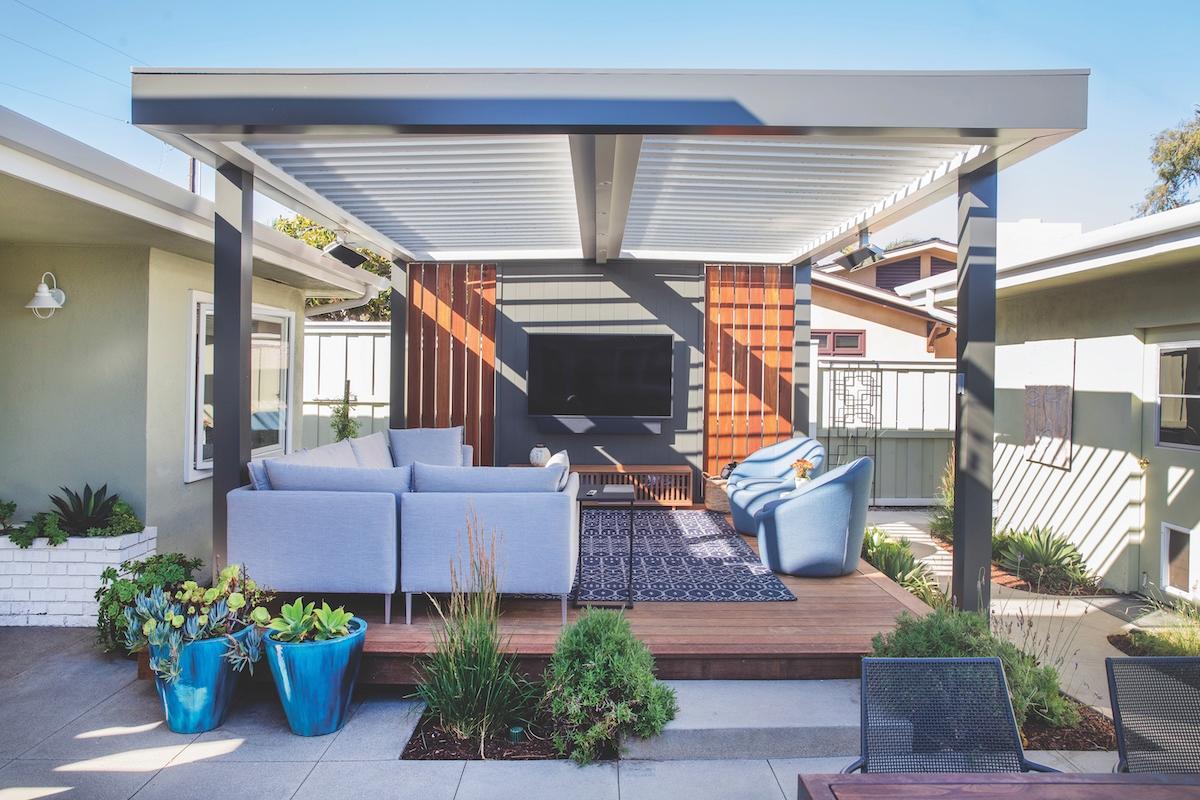 Contemporary backyard pergola with slatted roof, featuring an outdoor living room setup with sofa seating, lounge chairs, a mounted TV, and landscaped planters.