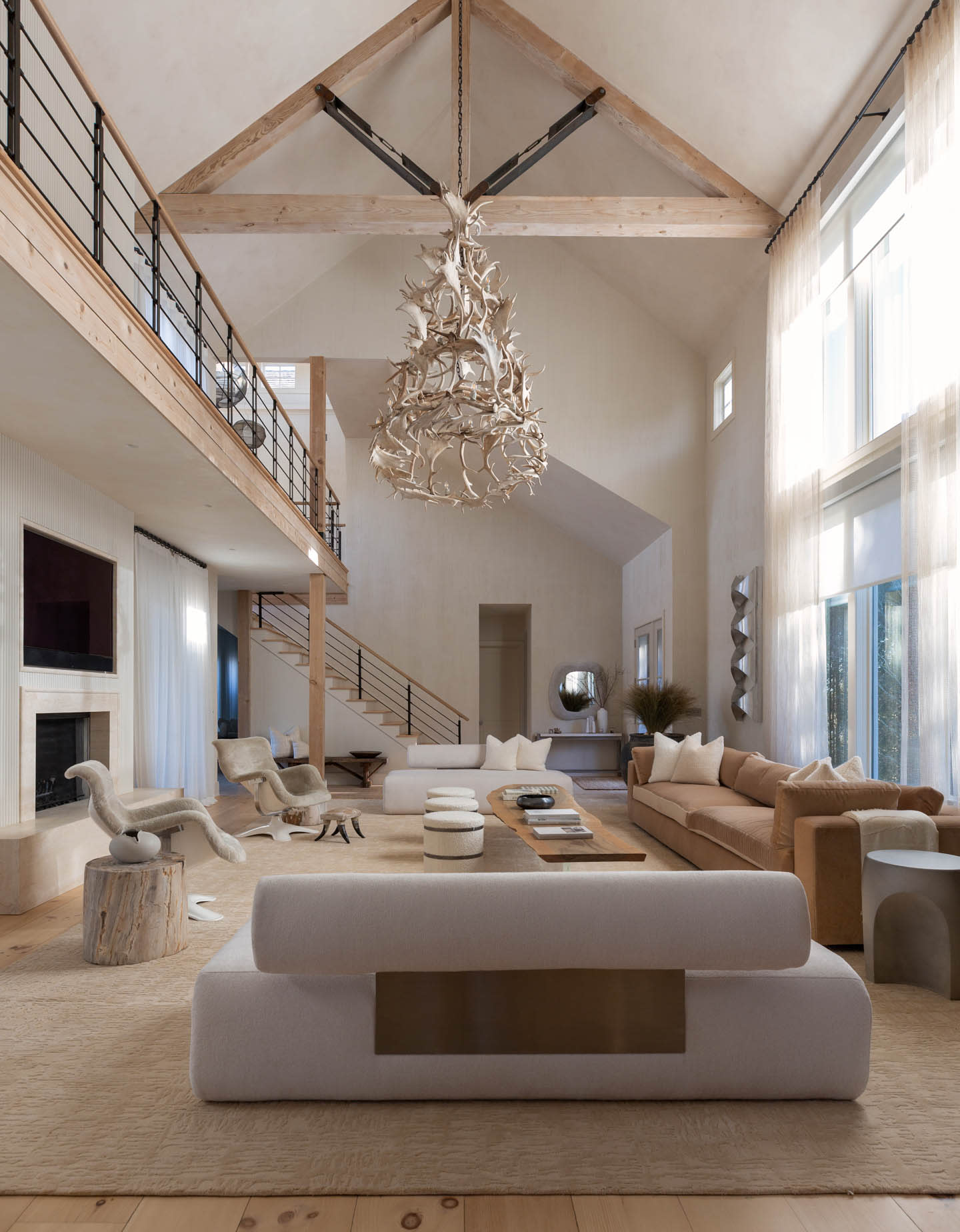 Spacious living room with high ceilings, a wooden beam, and a large antler chandelier. Neutral sofas, modern chairs, and large windows enhance the airy feel.