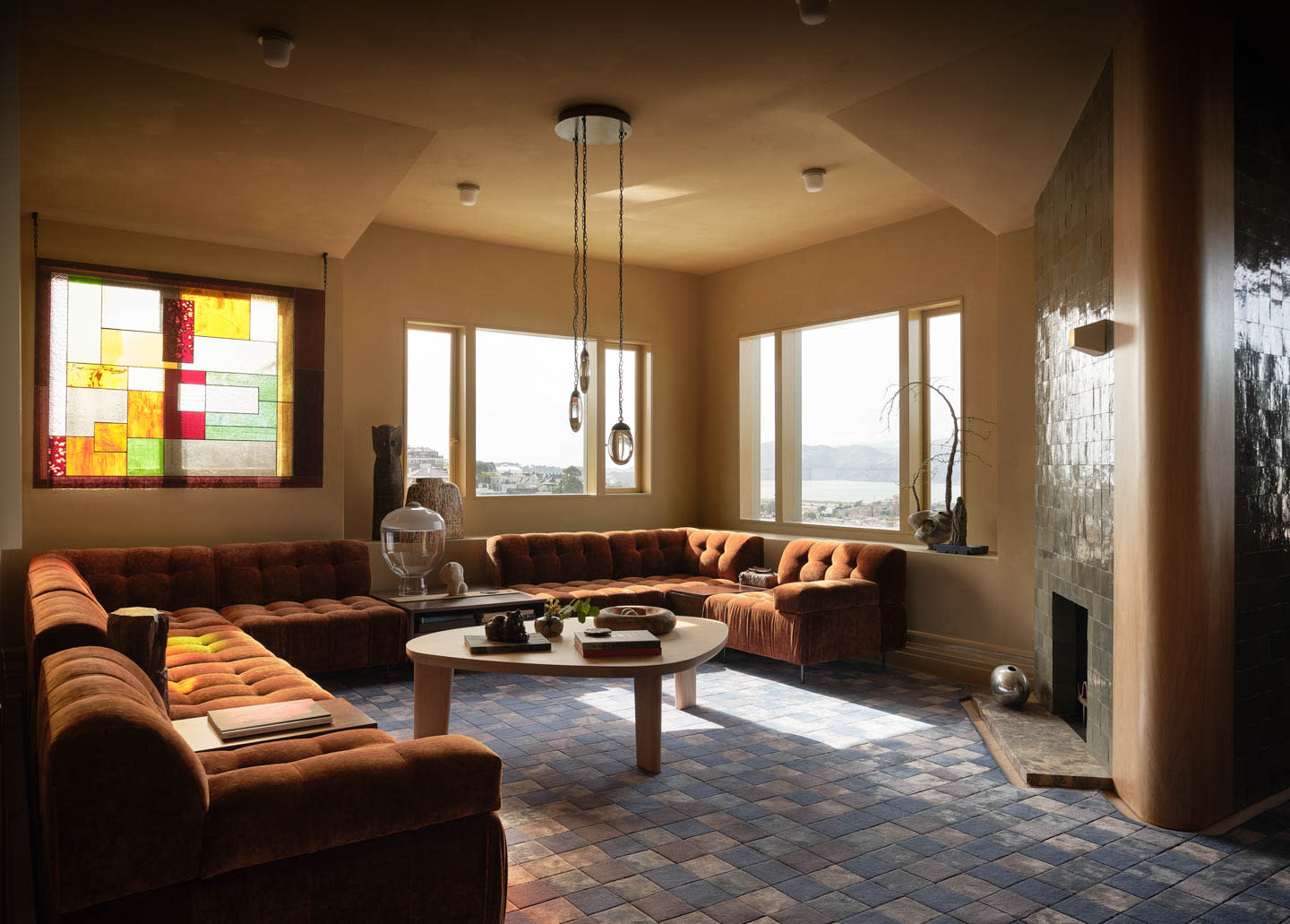 A cozy living room with a U-shaped brown velvet sofa, a round white coffee table, and large windows. A colorful stained glass panel adds warm light.