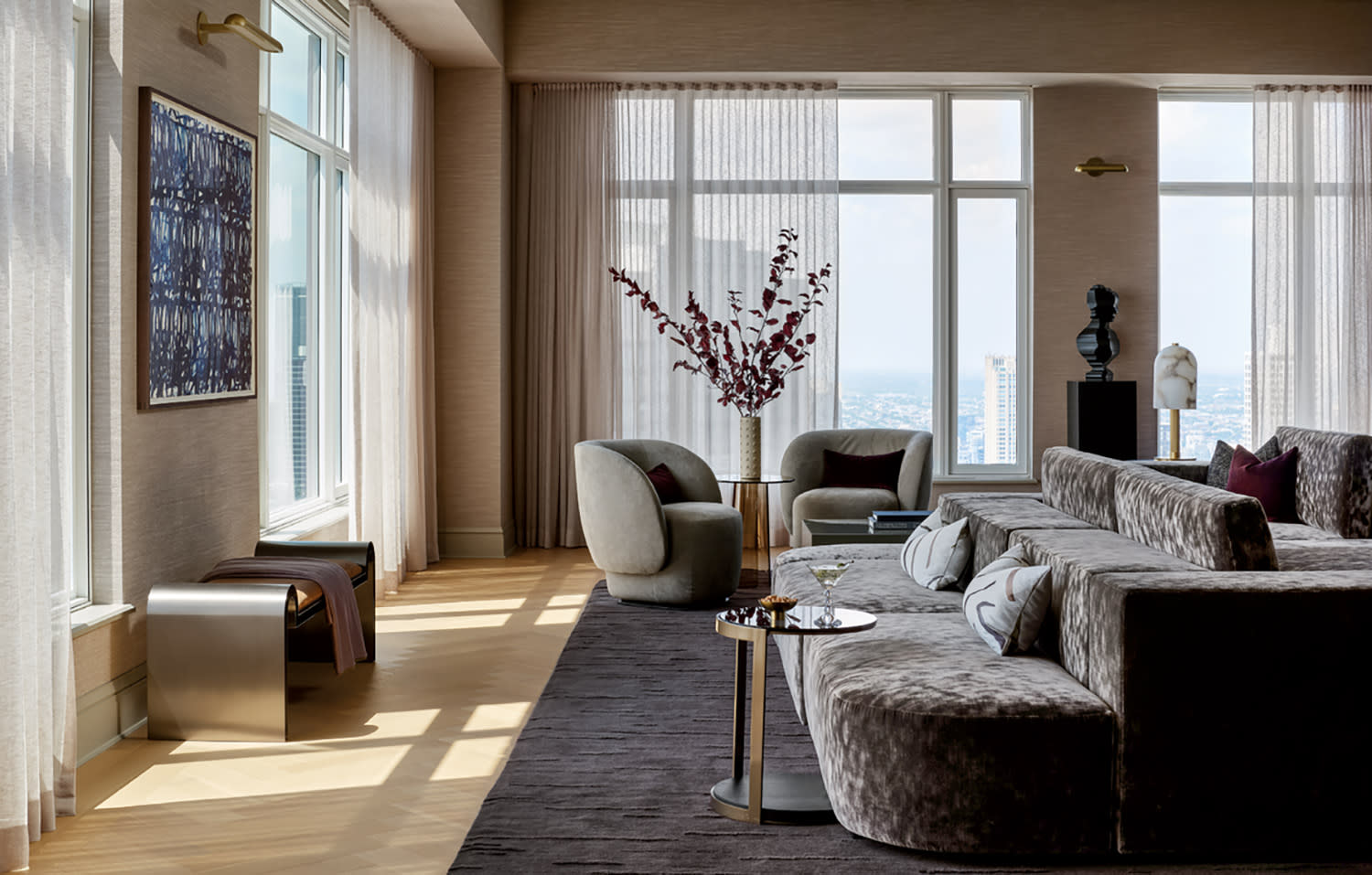 A luxurious living room with large windows, sheer curtains, and a city view. Features plush sofas, elegant armchairs, and a decorative vase on a table.