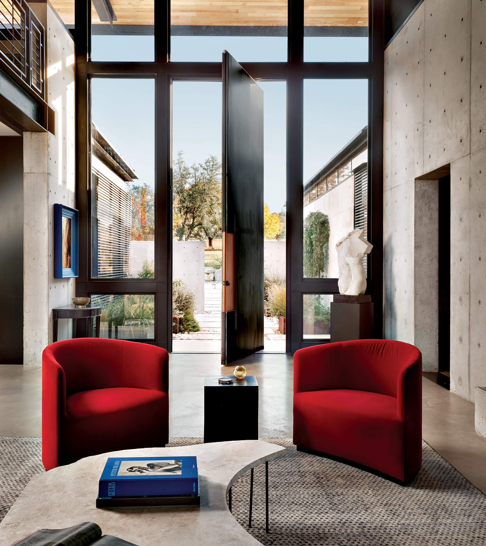 dramatic entry of a Brutalist home with a tall steel pivot door and red armchairs