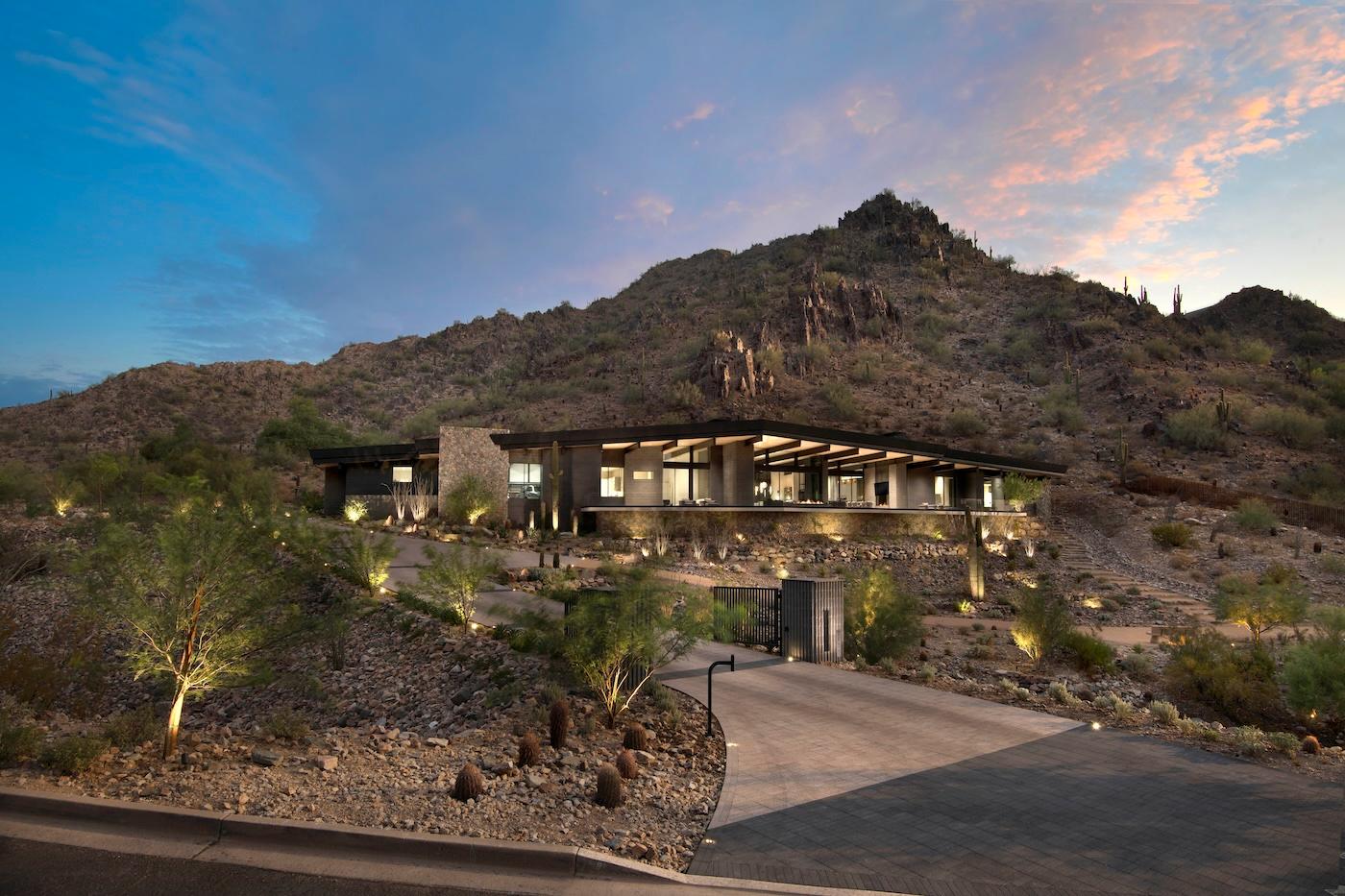 Modern hillside home with large windows, surrounded by desert landscape and cacti. Mountain backdrop at sunset, creating a serene and warm atmosphere.