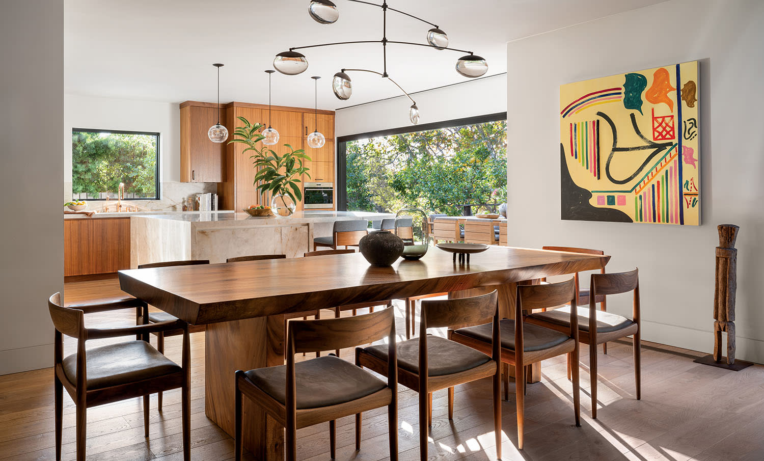 Modern dining room with a wooden table, six chairs, abstract art on the wall, and a large window offering a garden view. Warm, inviting atmosphere.