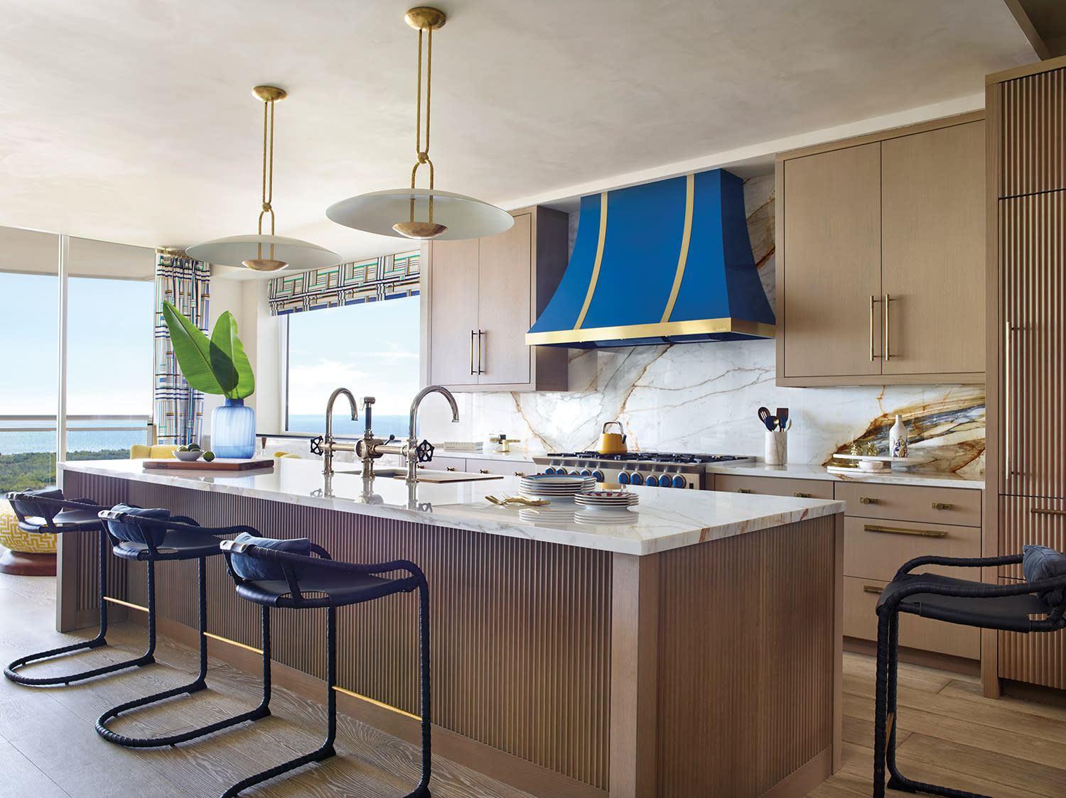 Modern kitchen with wooden cabinets, marble countertops, and a striking blue and gold range hood. Three sleek barstools and large windows offer ocean views.