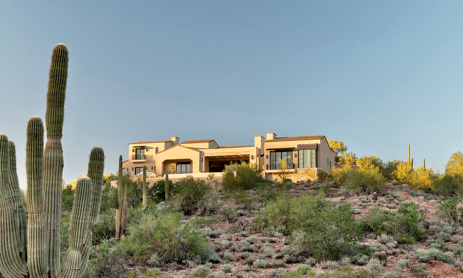 Desert home with beige stucco exterior on a hilltop, surrounded by cacti and sparse vegetation under a clear blue sky at sunset. Tranquil and remote.