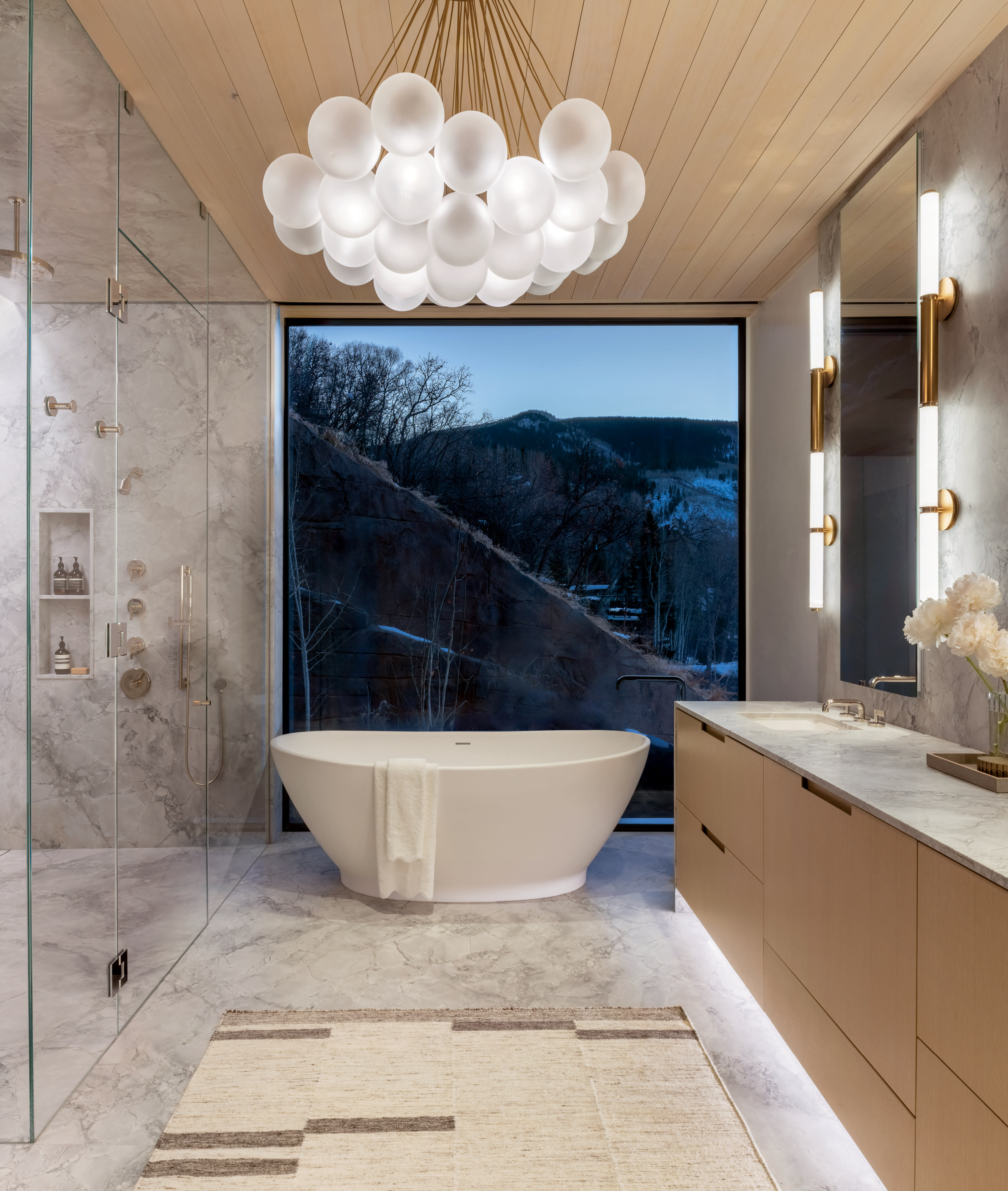 Modern bathroom with a freestanding bathtub, glass shower, and large window displaying a mountain view. Elegant lighting, marble surfaces, serene ambiance.