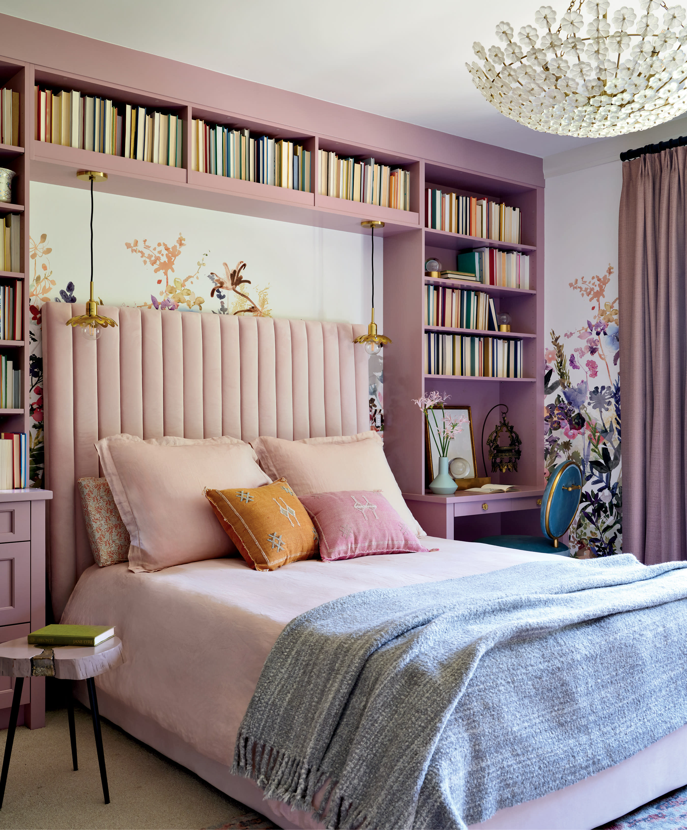 A cozy bedroom features a pink bed with colorful pillows and a gray throw. A floral mural and bookshelves surround it. A crystal chandelier hangs above.